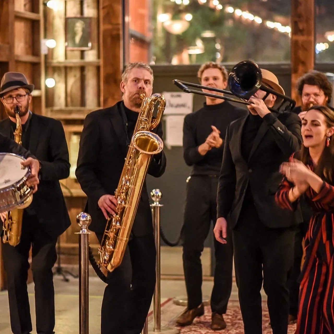 Musikere som spiller i et innendørs arrangement med treblåseinstrumenter, blant annet saksofon og trombone, med en kvinne som synger. Det er lyst og varm stemning, og publikum er til stede.