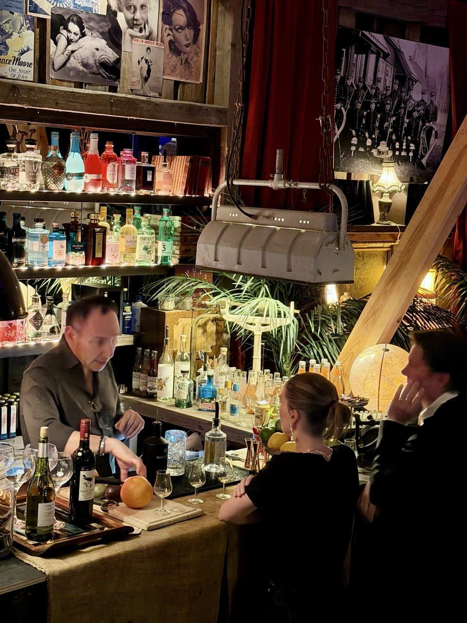 En bartender serverer drinker på en bar, mens to kunder ser på. Baren er fylt med ulike typer alkoholflasker og dekorasjoner, og bakgrunnen har bilder og plakater på veggen.
