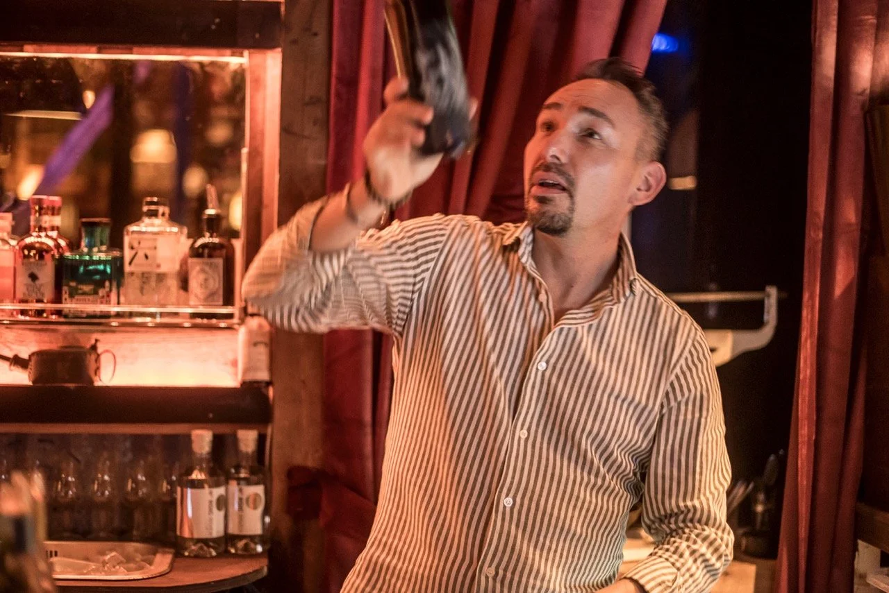 En mann som jobber som bartender i en bar, skjenker en drink, omgitt av alkoholflasker.