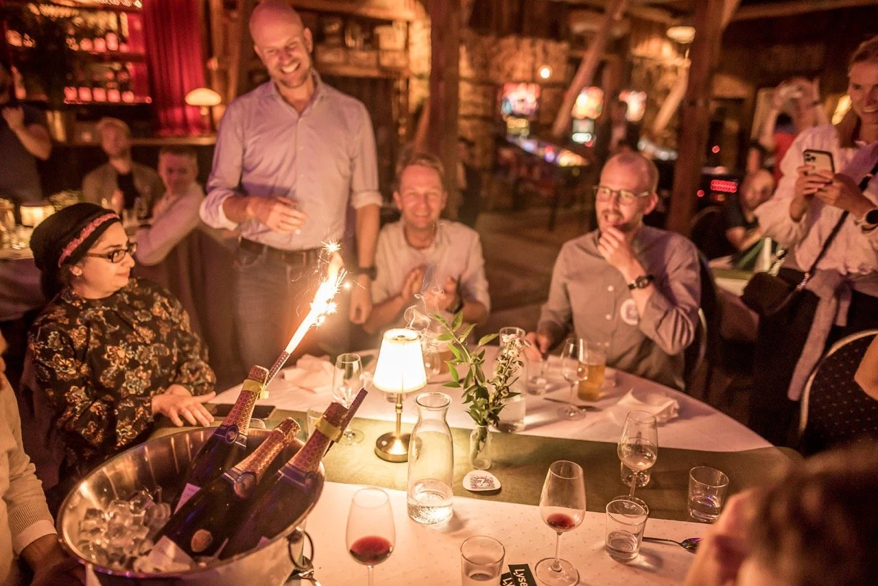En gruppe mennesker feirer bursdag med champagne og bållys på et bord, i en rustikk varm atmosfære i en restaurant eller bar.