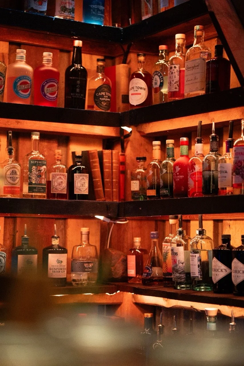 Hylla med ulike typer alkoholholdige drikker, inkludert gin og likører, mot en trevegg med varmt belysning.