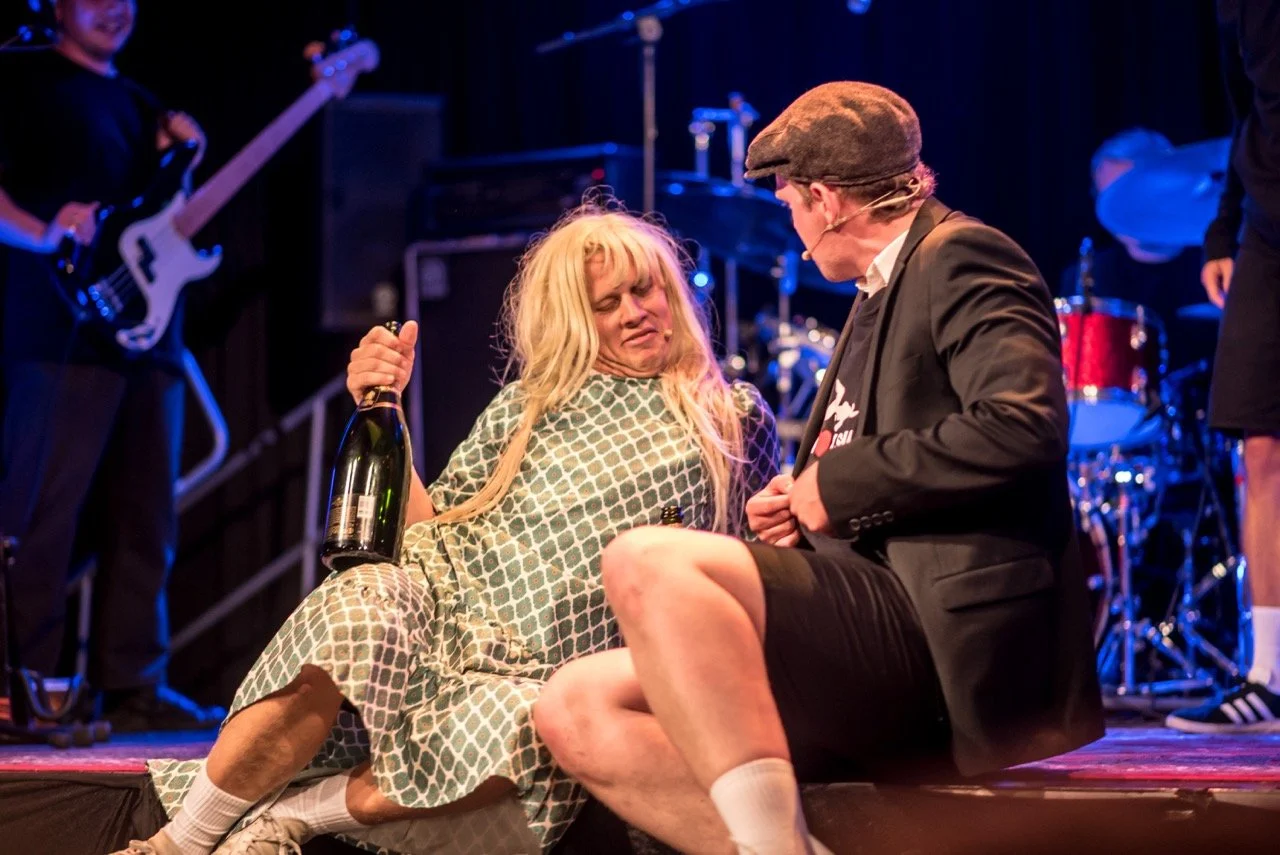 To personer sitter på scenen under en forestilling, en kvinne med langt blondt hår og en mann med briller og flat cap. Kvinnen holder en flaske og ser ut til å ha vondt, mens mannen har på seg en svart dressjakke.