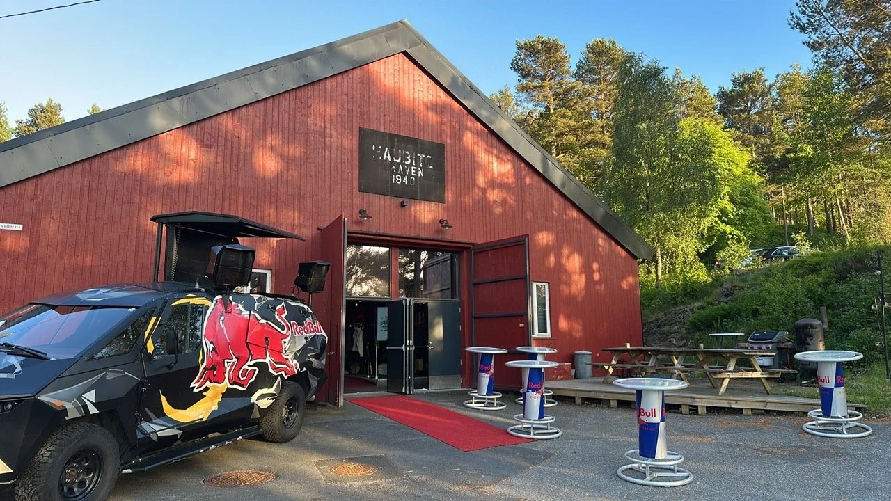 En rødt trebygning med åpen inngangsdør og en Red Bull-annonseringsbil parkert foran. Uteområde med bord og stoler, samt et grillskap i bakgrunnen. Natt og trær i bakgrunnen.