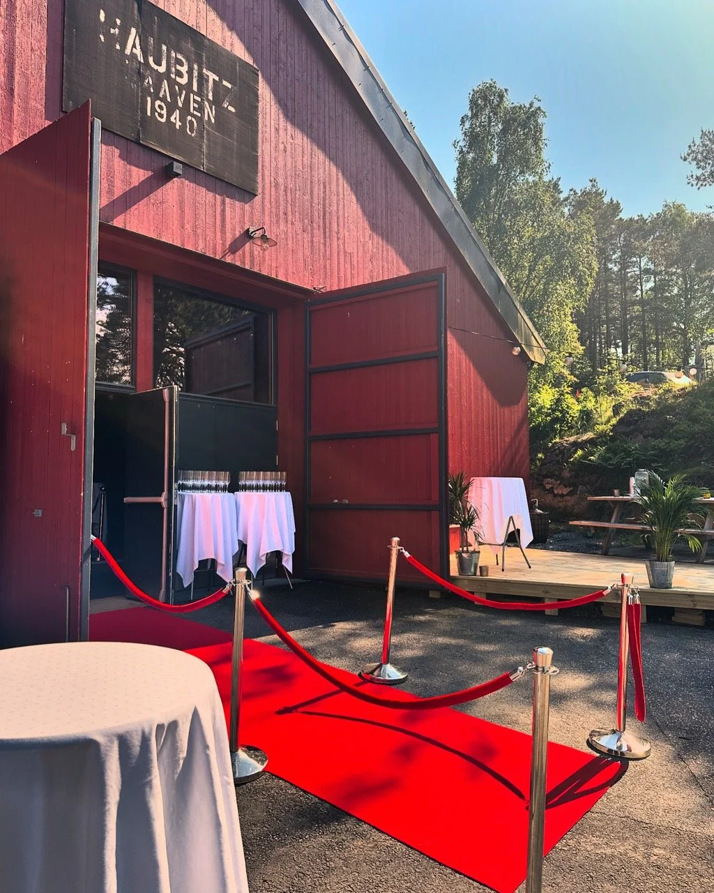 En rød teppe ruller ut foran en inngang til en bygg med røde vegger, dekorert for en spesiell anledning med stoler og planter utenfor. Pynten indikerer en formell begivenhet.