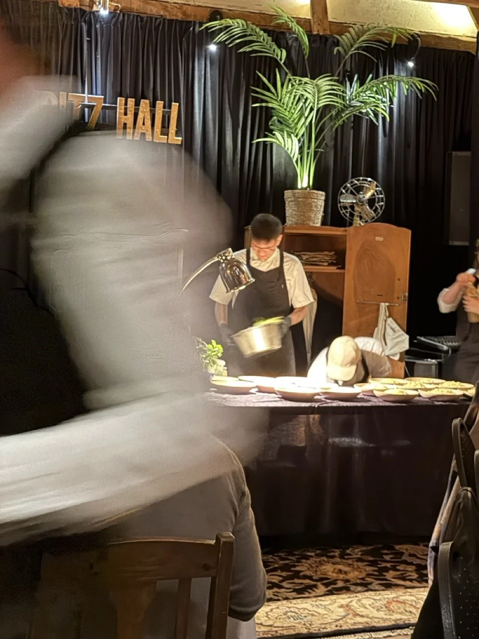 Et bilde av en innendørs scene med kokker som forbereder mat. En kokk i forgrunnen holder en metallbolle, mens en annen bøyer hodet over en borddekket mat. Bak dem er det en stor plante, en vifte, og en svart bakgrunn med et skilt som sier '7 HALL'.