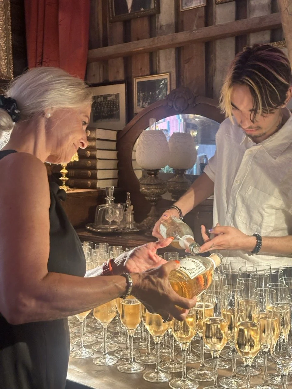 En eldre kvinne og en yngre mann som heller champagne i glass, i et rom med trevegger, bøker og gamle lamper.