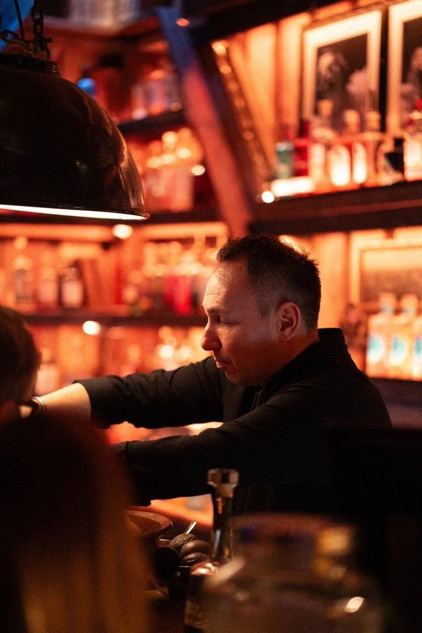 En mann i svart skjorte som jobber bak en bar, omgitt av brune trevegger og hyller med flasker og bilder, i en dimly lit bar eller pub.