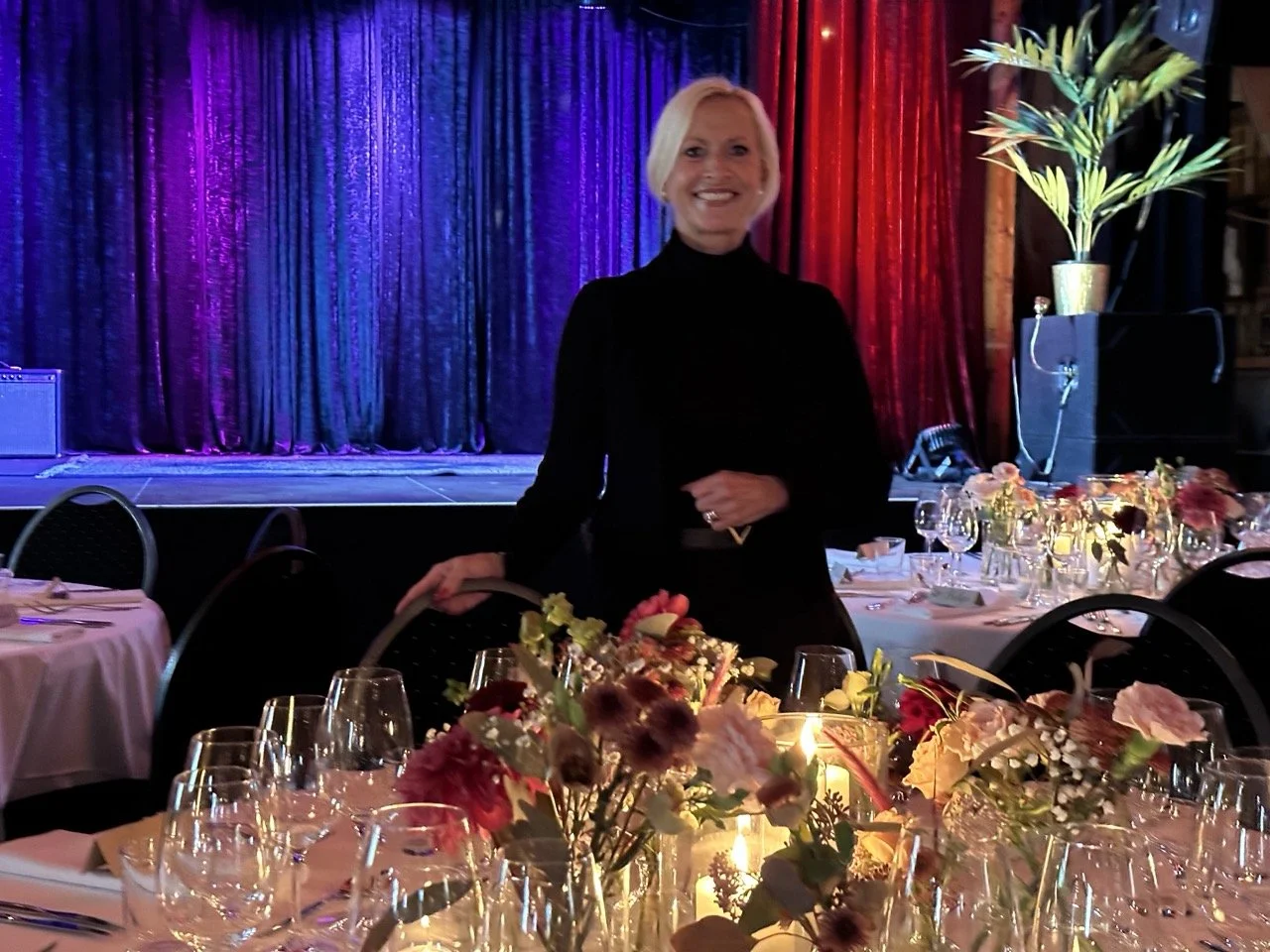 En smilende kvinne med blond hår i svart antrekk står foran en borddekorasjon med blomster og lys på et festbord. Bak henne er en scene med fargerike gardiner.