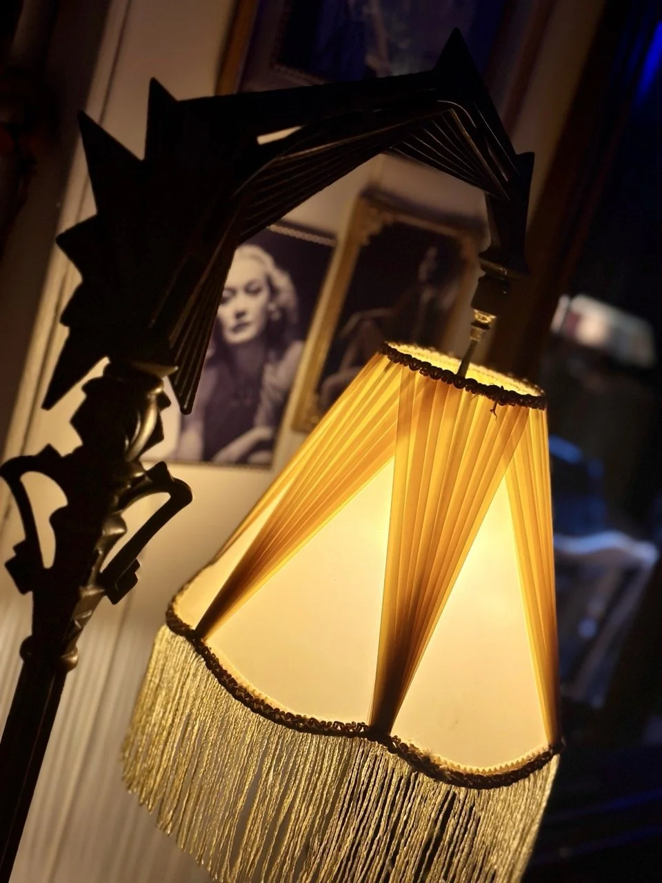 Et bordlampe med en gul, plettet skjerm og frynsede detaljer henger fra taket. I bakgrunnen er det bilder av kvinner på veggen.