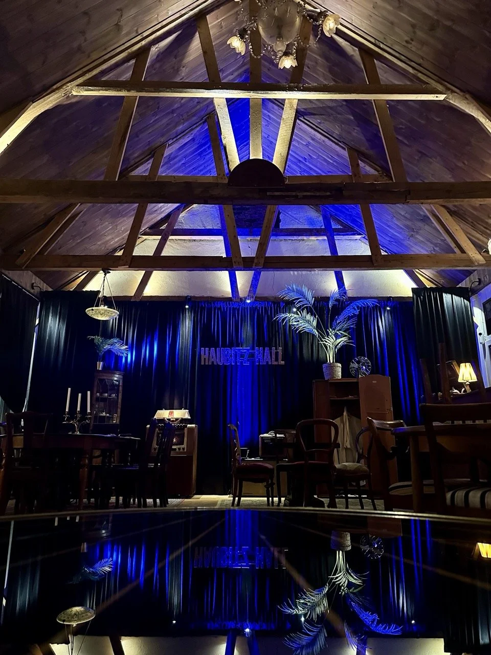 Interiør av et rom med høyt tak, treverk og stoler, en scene med en svart bakgrunn og lys, dekorative planter og lys donner. Teksten "HAUBITE HALL" er synlig på scenen.