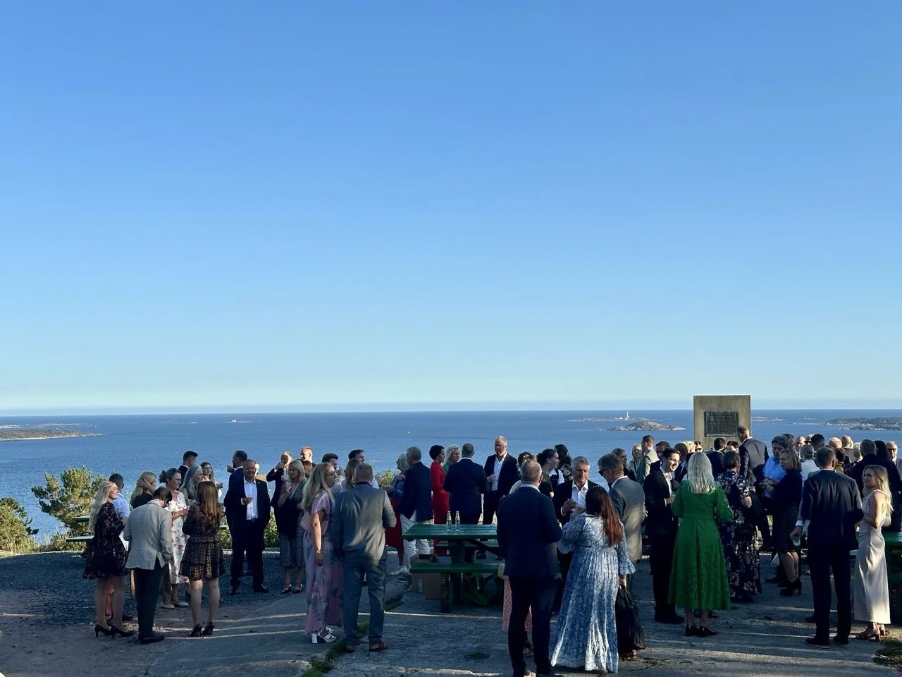 En gruppe mennesker i formelle klær samles utendørs foran en havutsikt under en solrik dag.