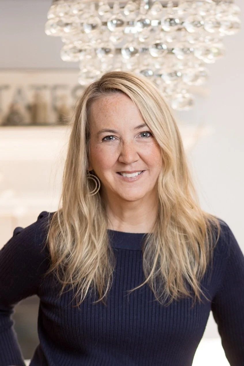 Smiling woman with long blonde hair wearing a navy sweater, standing indoors with a decorative light fixture in the background.