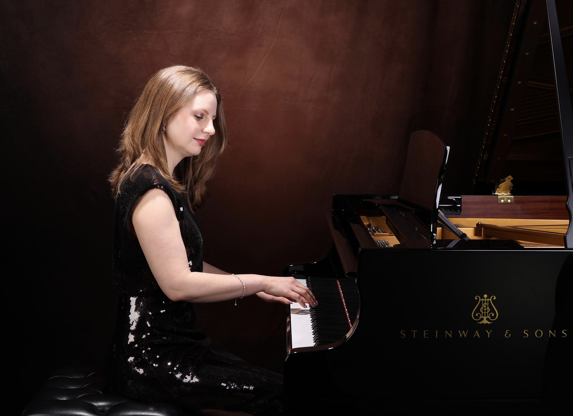 A woman with light skin and shoulder-length brown hair playing a black Steinway & Sons grand piano in a dimly lit room.