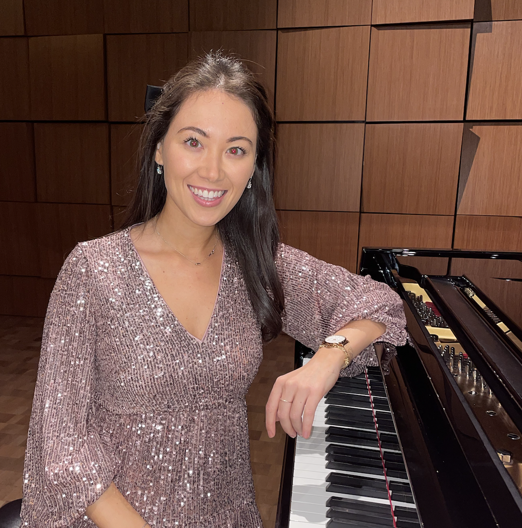A smiling woman with long dark hair wearing a sparkly pink dress, sitting at a grand piano in a room with wooden paneled walls.