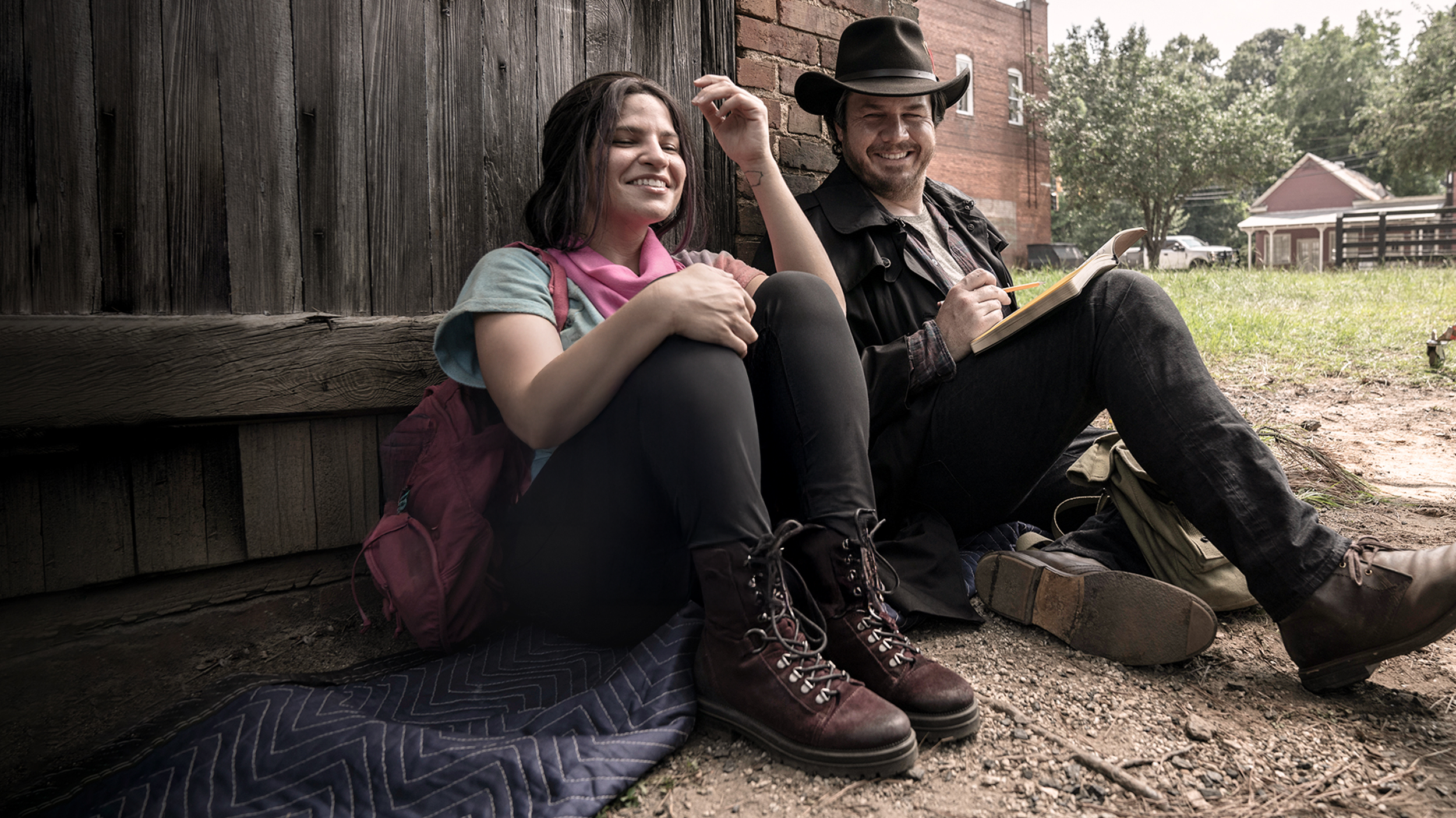 Two survivors relaxing outside a rustic building, sharing a light moment in “The Walking Dead”