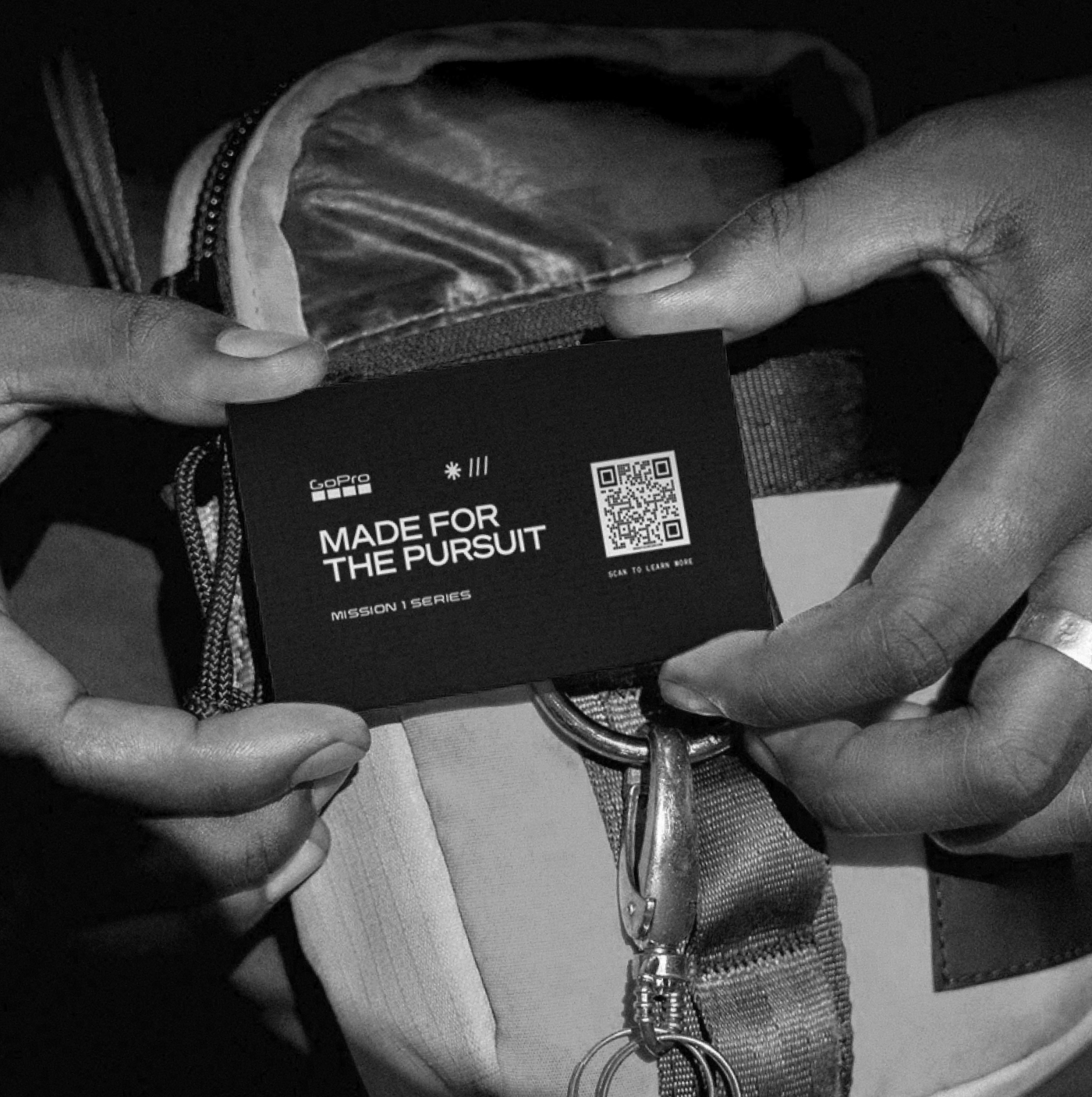 Hands holding a tag that says 'Made for the Pursuit' with a QR code and the GoPro logo, attached to a backpack with a strap and zipper.