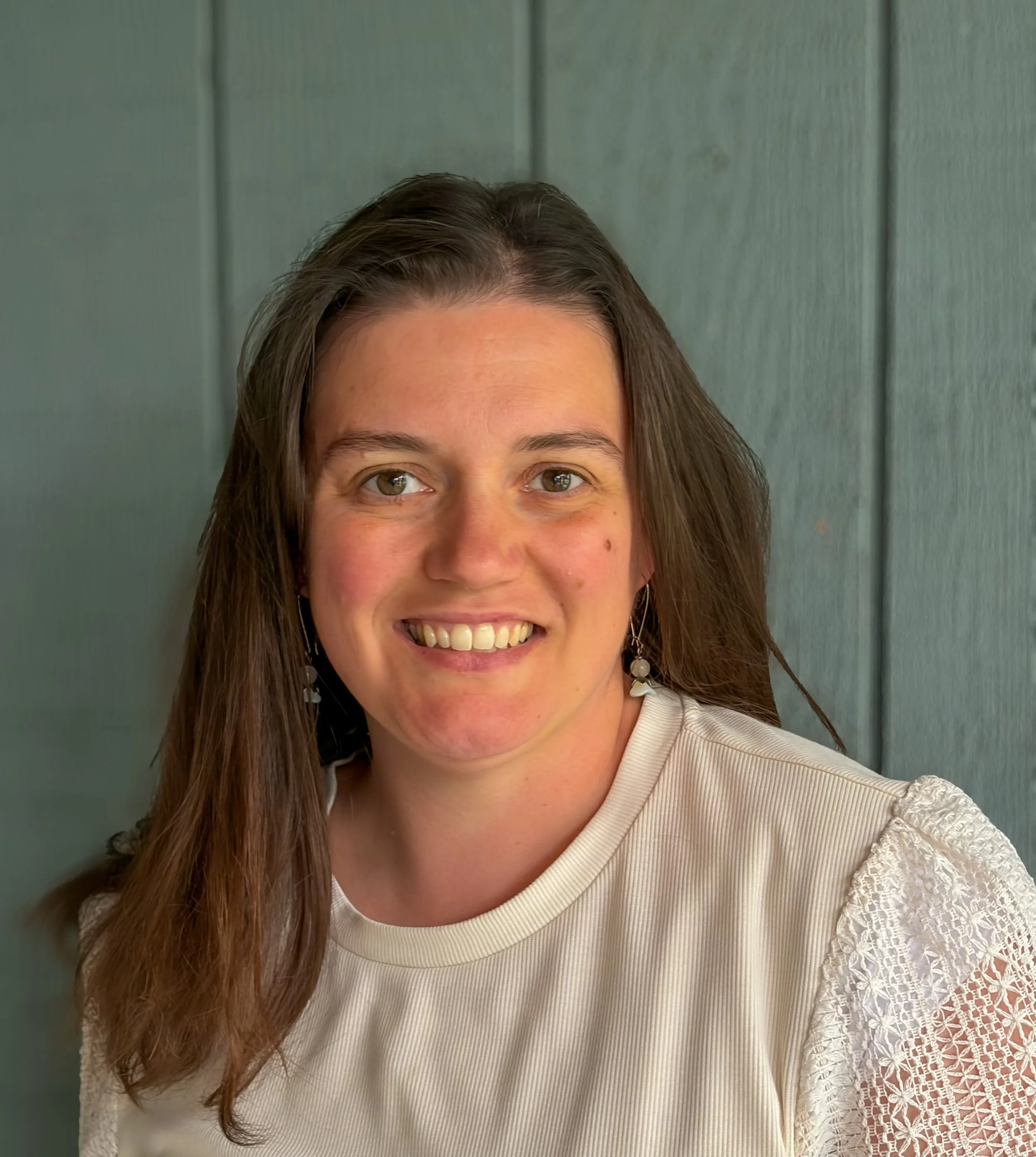 Close-up of a woman with brown hair wearing a cream-colored top with lace details on the sleeves, smiling and looking at the camera, standing against a gray paneled wall.