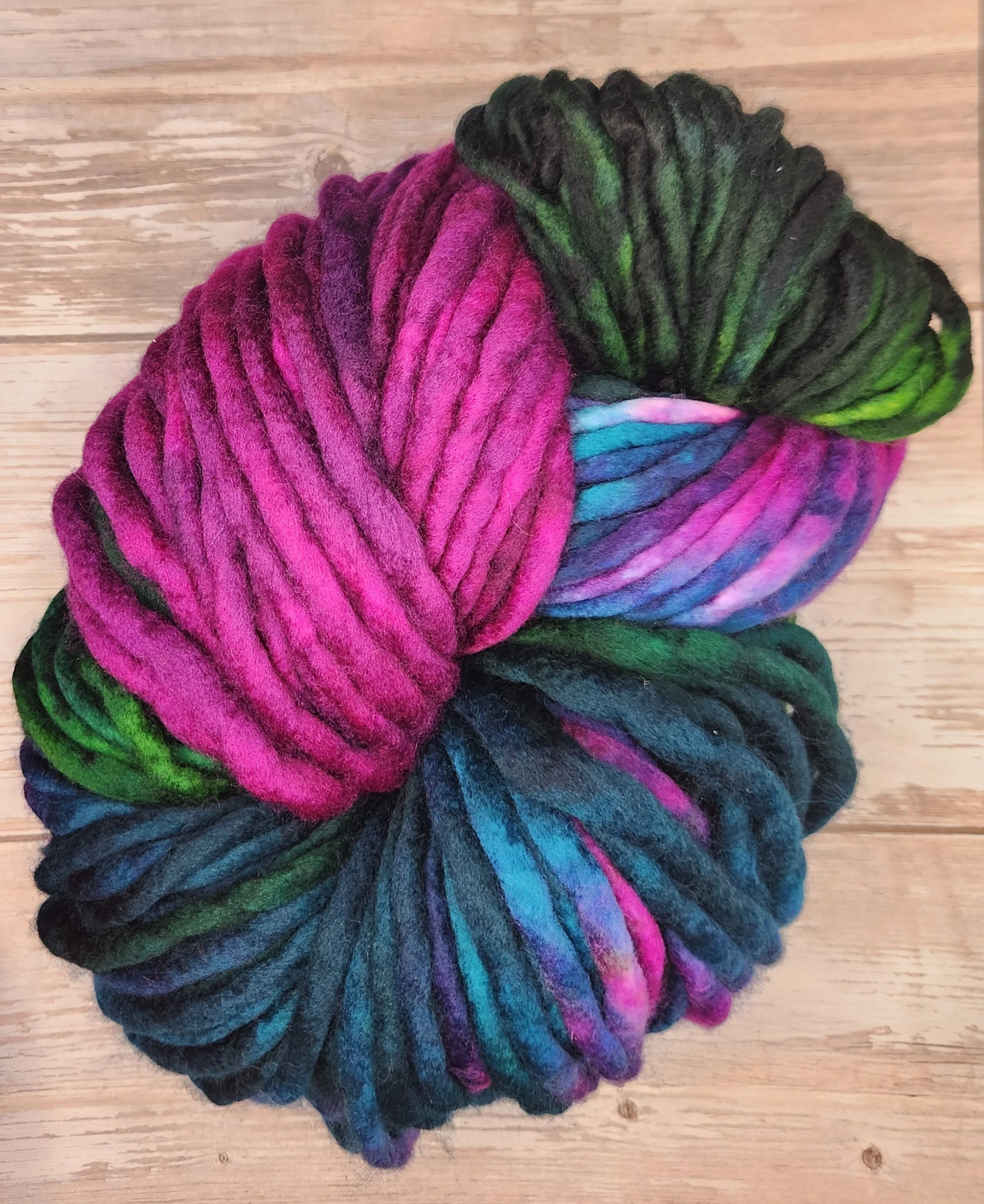 Colorful yarn skeins in pink, green, black, and multicolor shades, arranged in a circular pattern on a wooden surface.