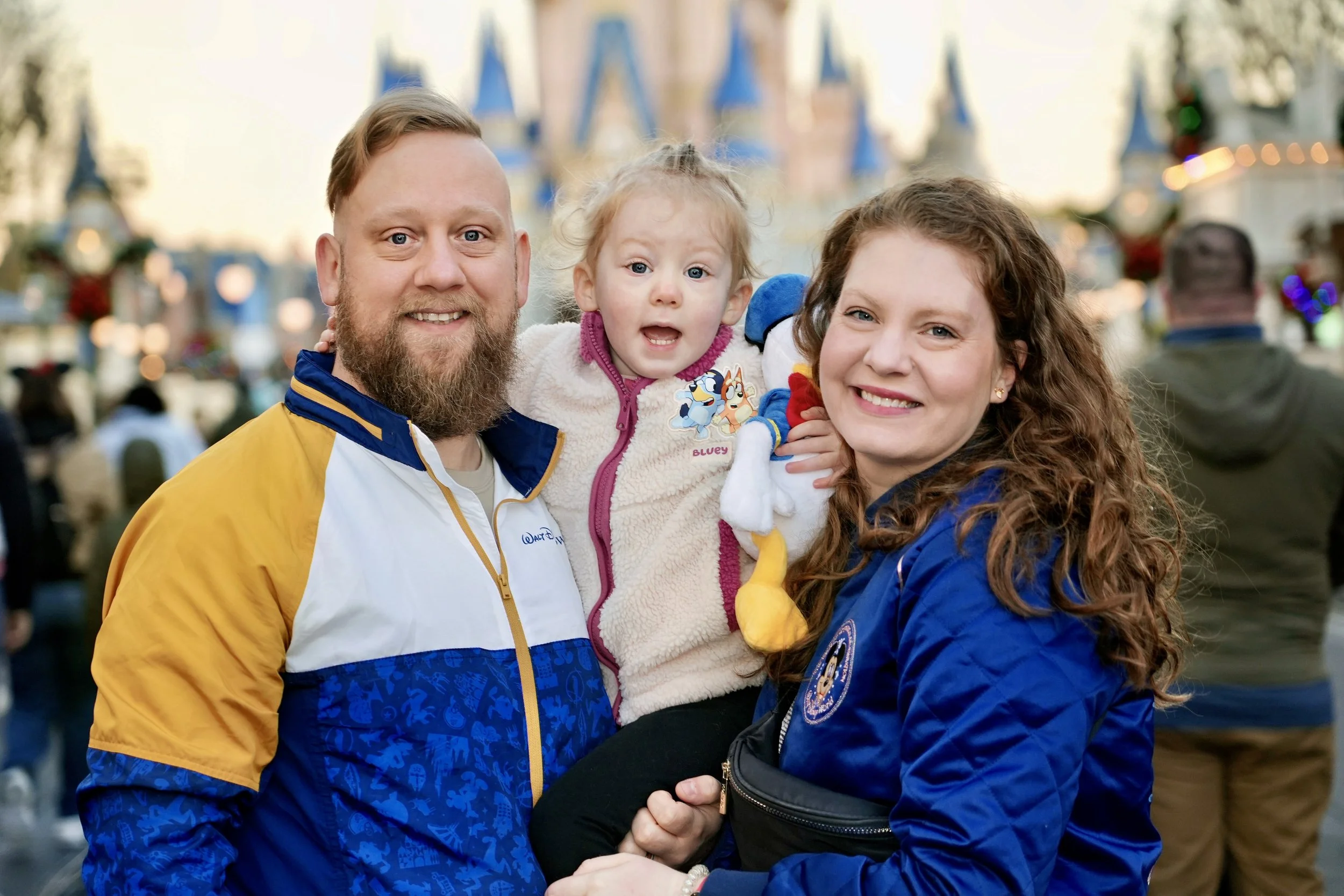 A family of three smiling and posing at a theme park, with a castle in the background. The father has a beard and wears a blue and yellow jacket, the mother has curly hair and wears a blue jacket, and the young girl is held between them, wearing a fleece jacket with cartoon characters.