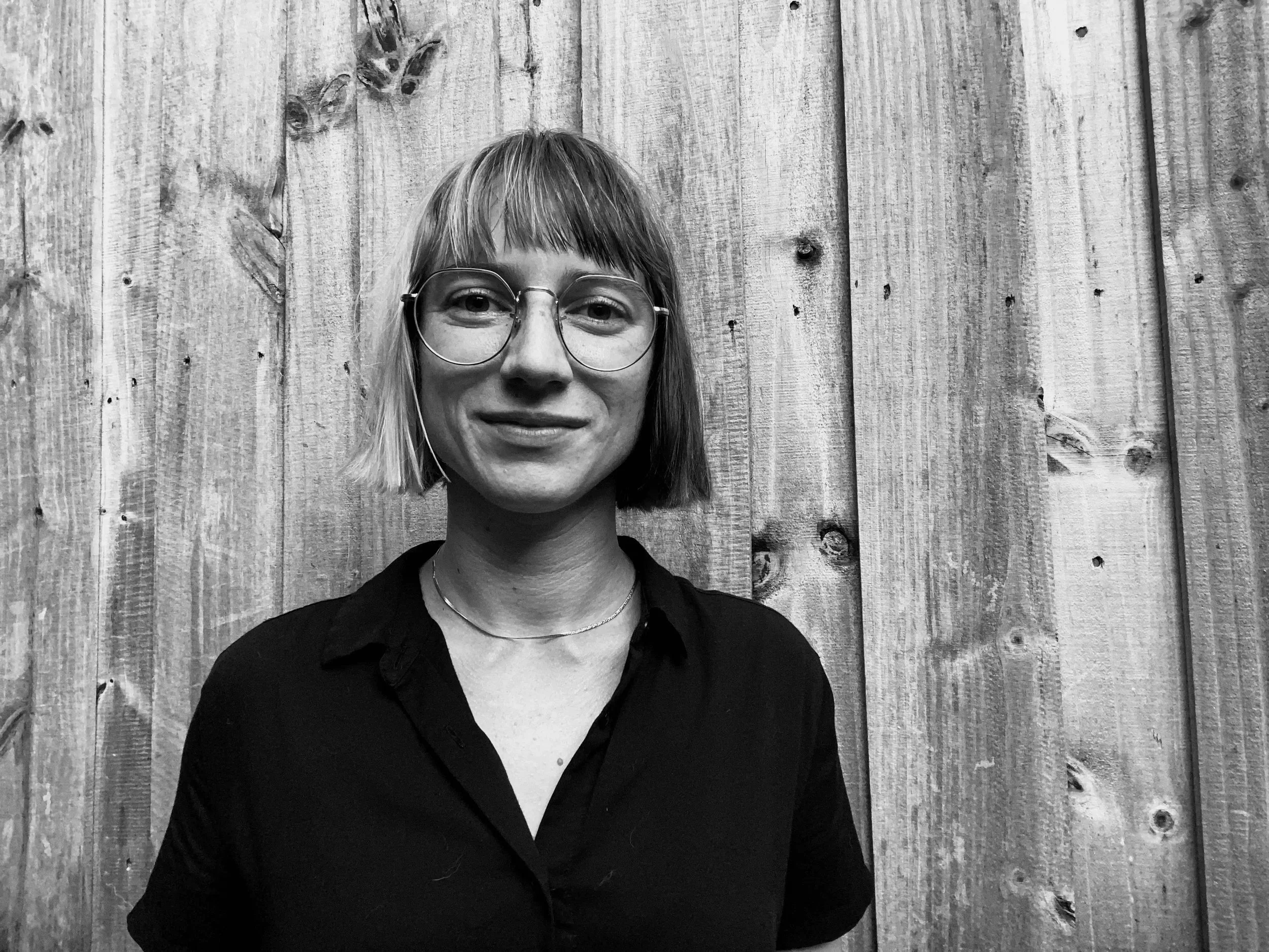 Black and white photo of a woman with short hair and glasses, smiling in front of a wooden wall.