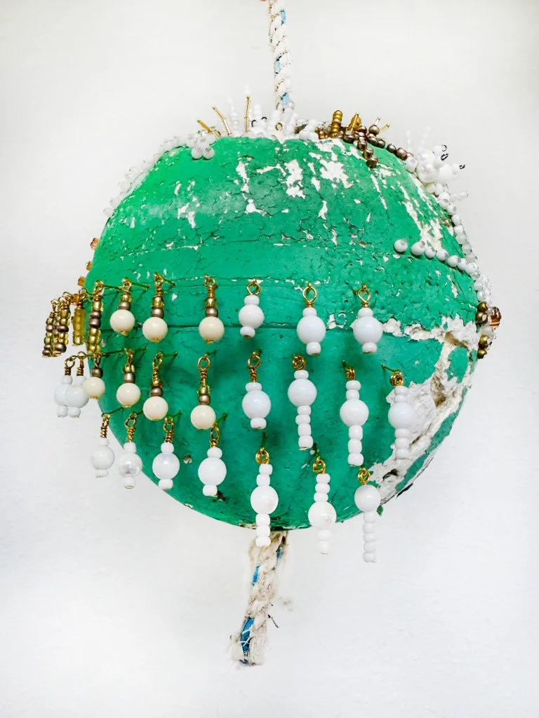 A green spherical object with white beaded jewelry and ornaments hanging on it, attached to a white background by a frayed rope.