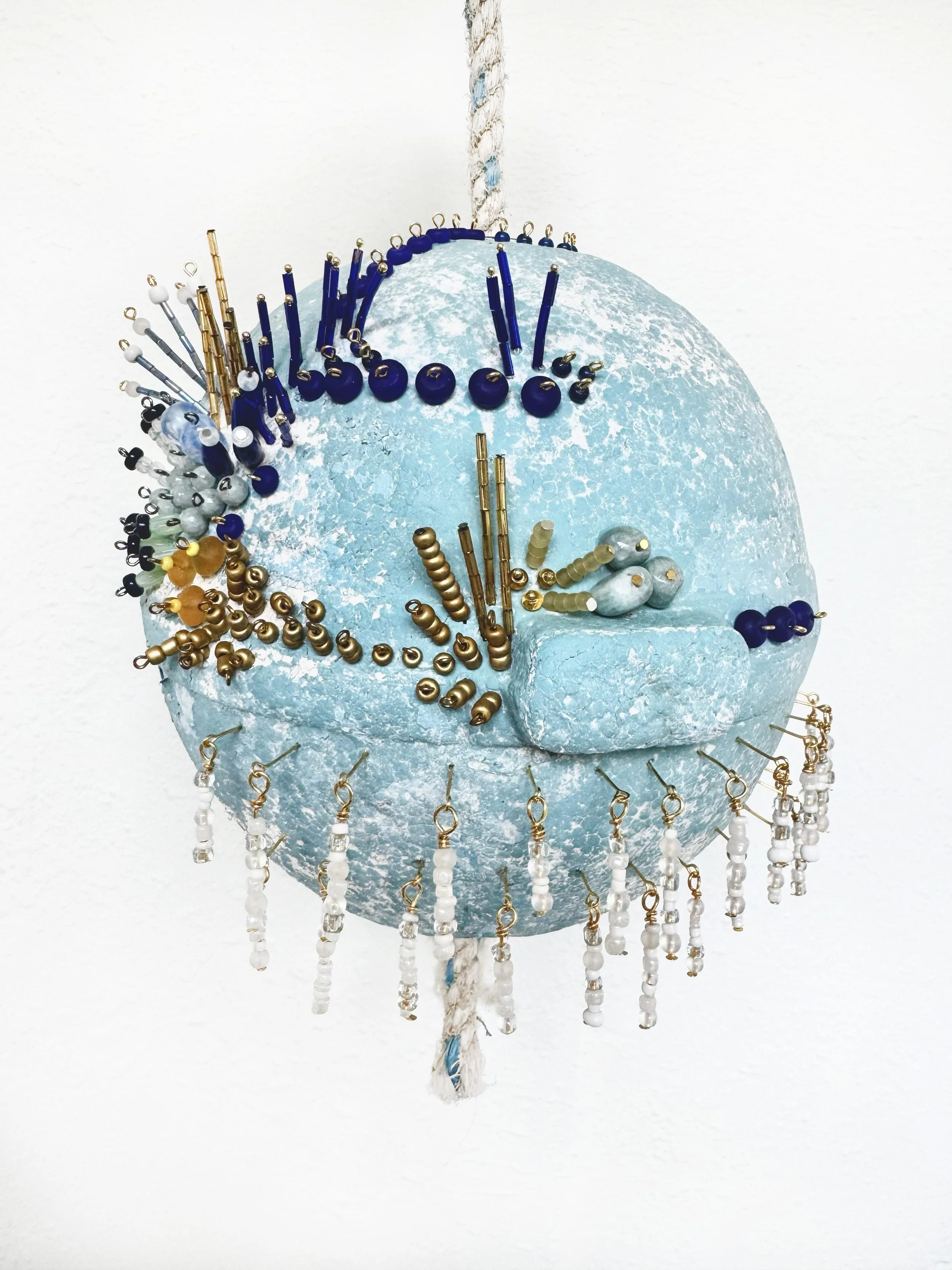 Decorative clock sculpture made of a round, light blue textured material adorned with various categorized beads and small hanging beads, suspended by a white twisted rope.
