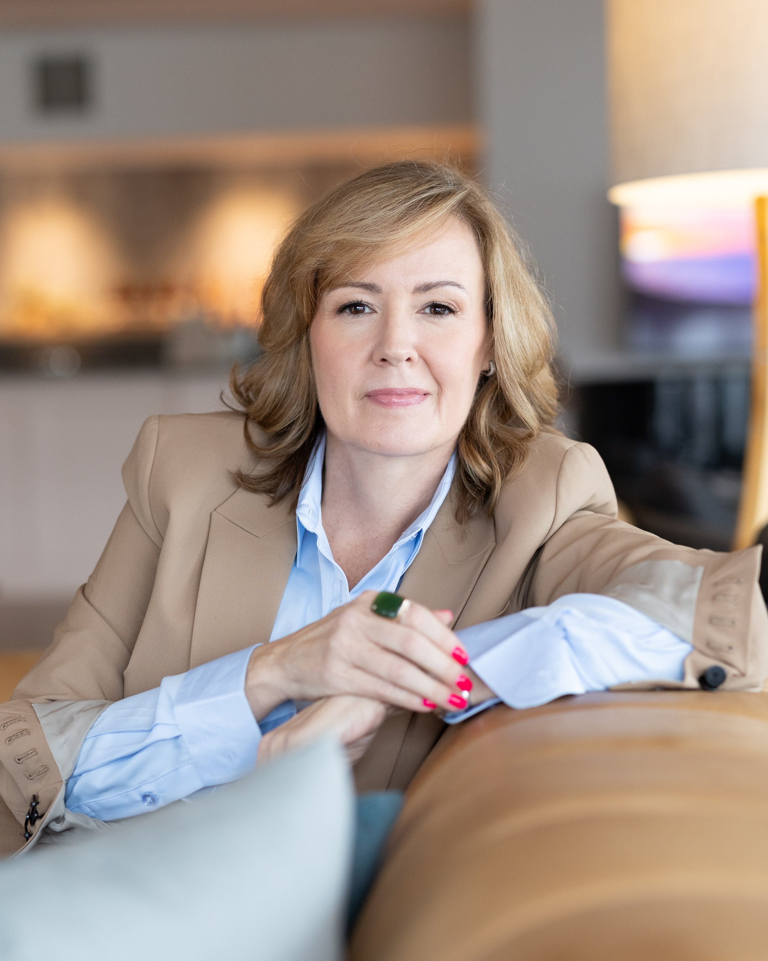 A woman with blonde hair, wearing a beige blazer and a blue shirt, is smiling and holding a pen or marker, possibly during a meeting or discussion in an office setting with blurred background lights.