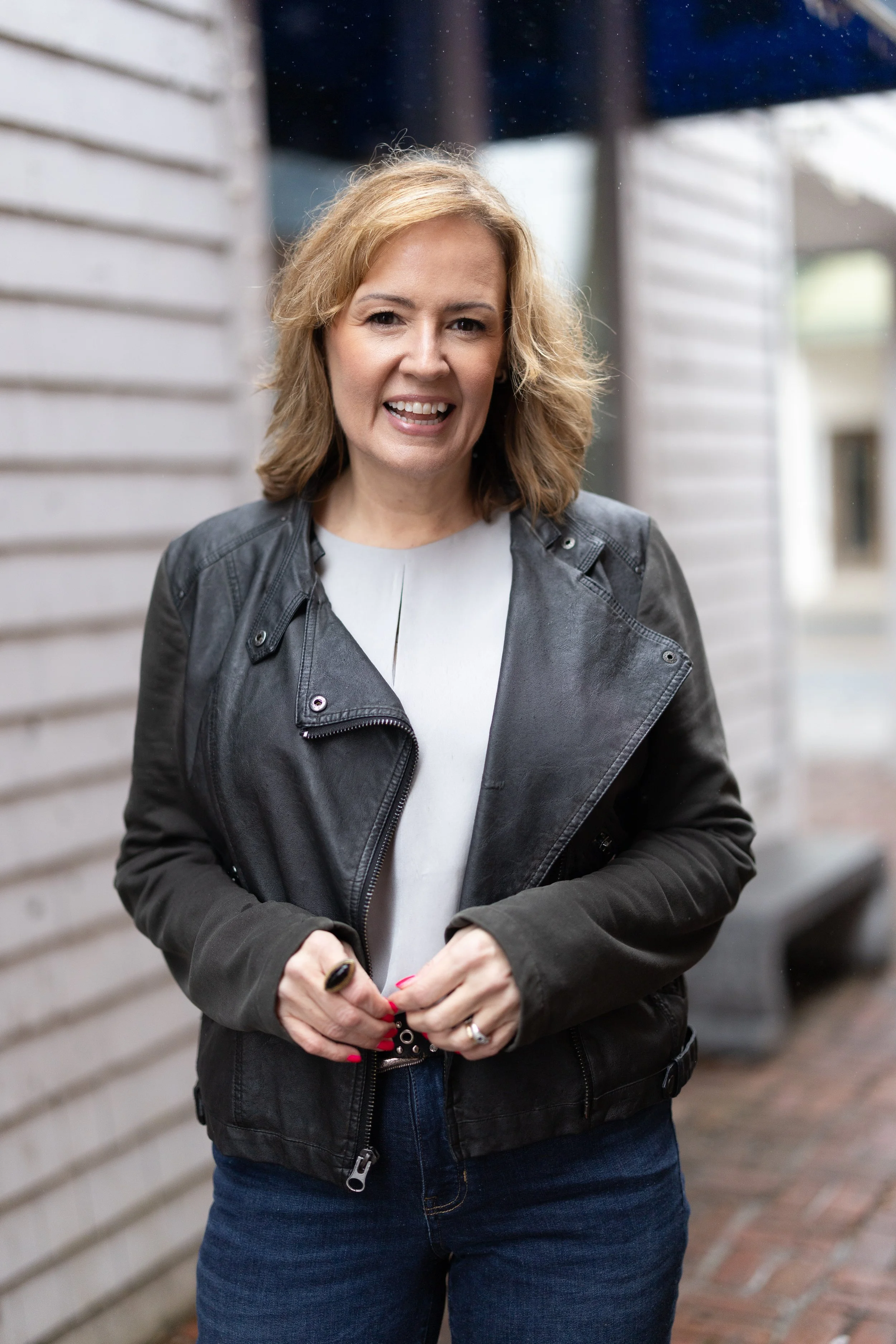 Women with blonde hair wearing a black jacket and jeans standing outdoors against a wooden siding wall.