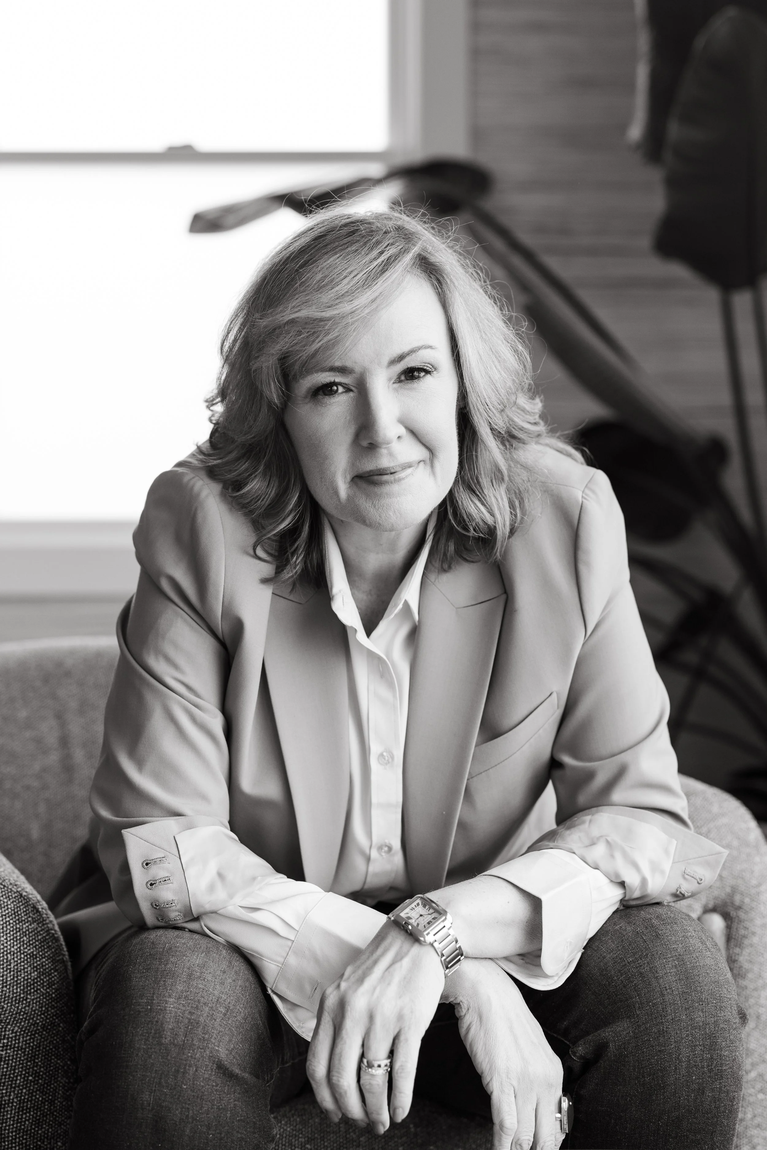 Black and white photo of a woman with shoulder-length hair, smiling, sitting on a sofa with her arms resting on her knees. She is wearing a blazer, a white shirt, a watch, and rings. The background includes a window and some clothing hanging on a wooden rack.