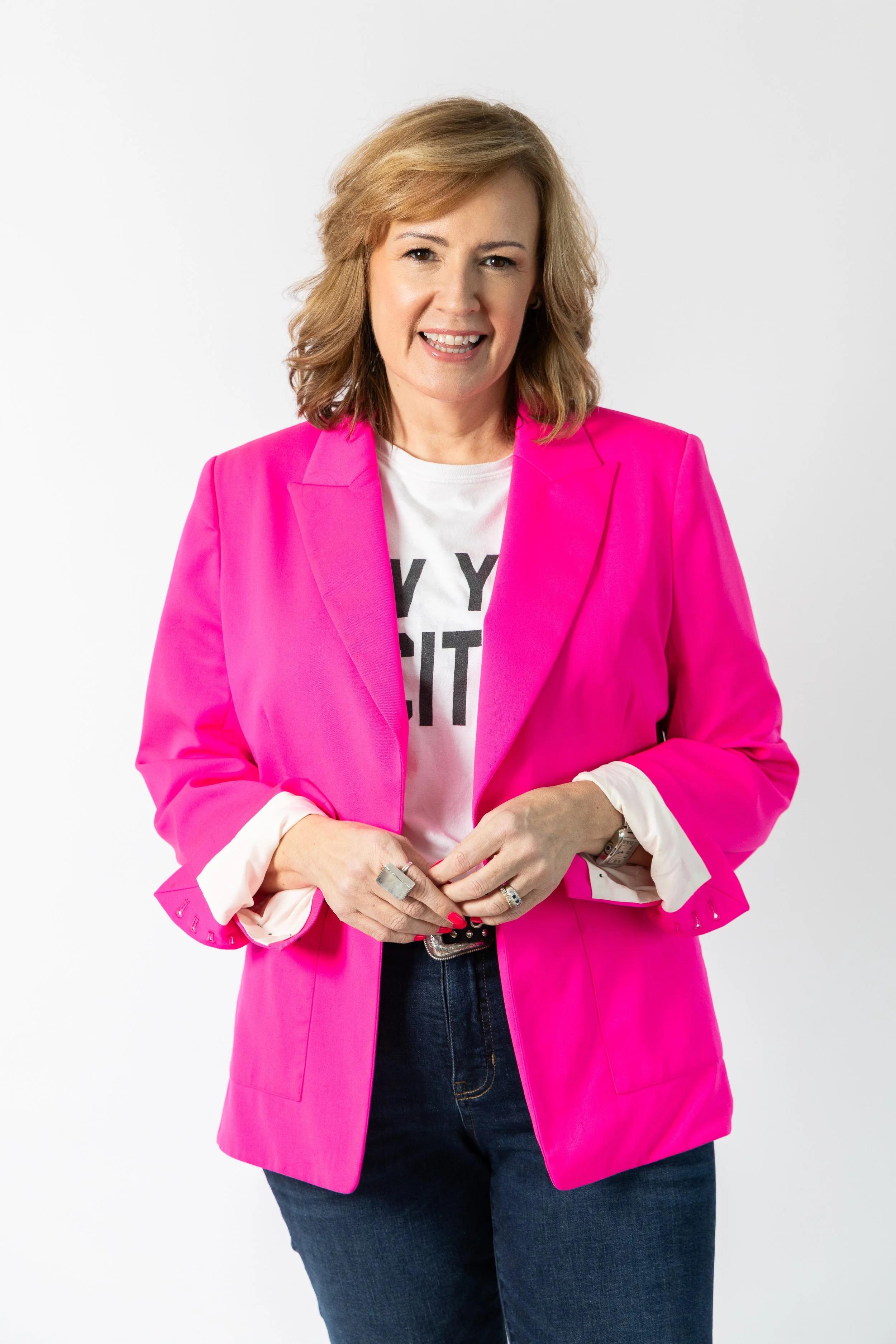 Woman with shoulder-length hair wearing a bright pink blazer over a white graphic T-shirt, dark jeans, and rings on fingers, standing against a plain white background.
