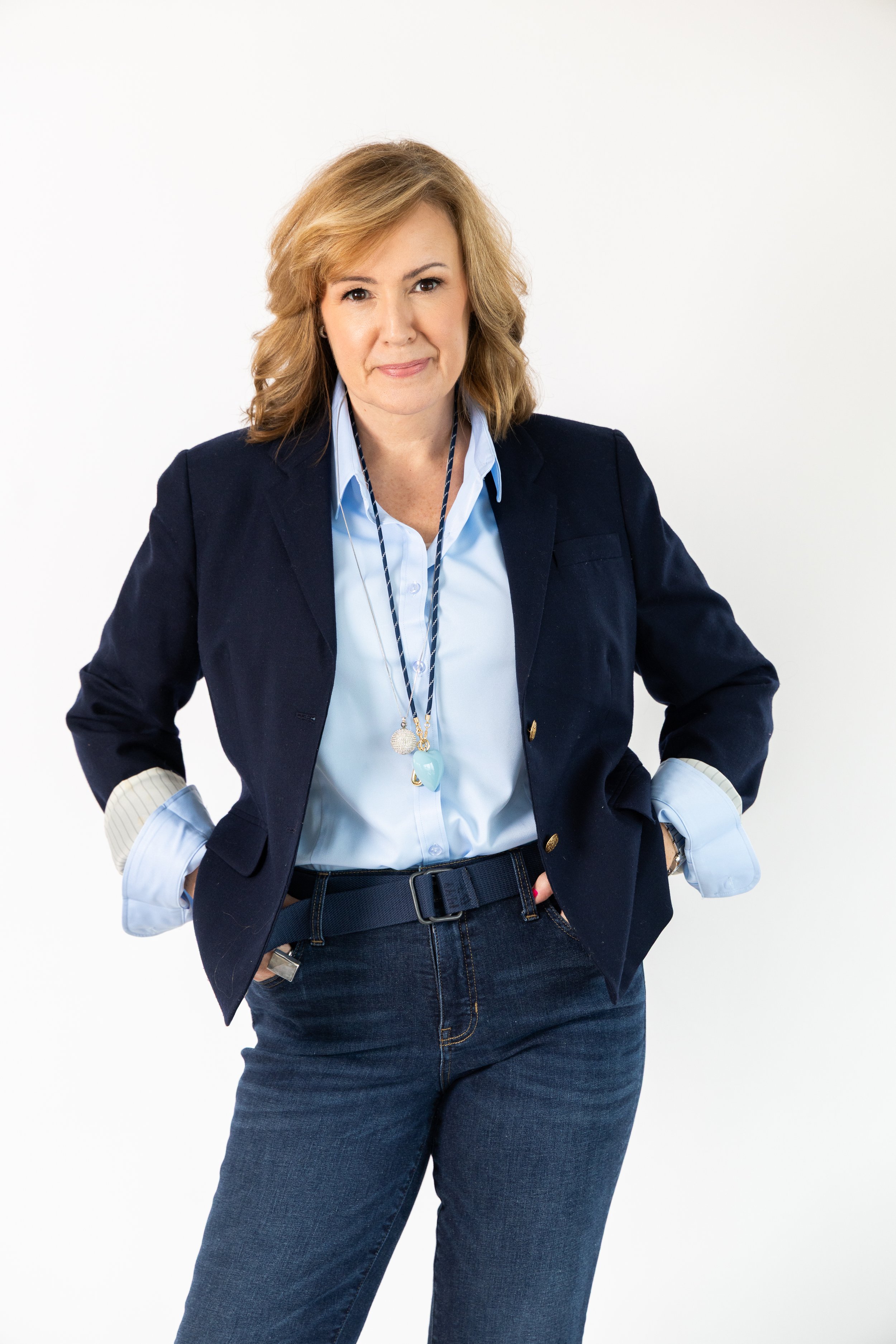 A woman with light brown hair, wearing a dark blazer over a light blue shirt, standing with her hands in her pockets against a white background.