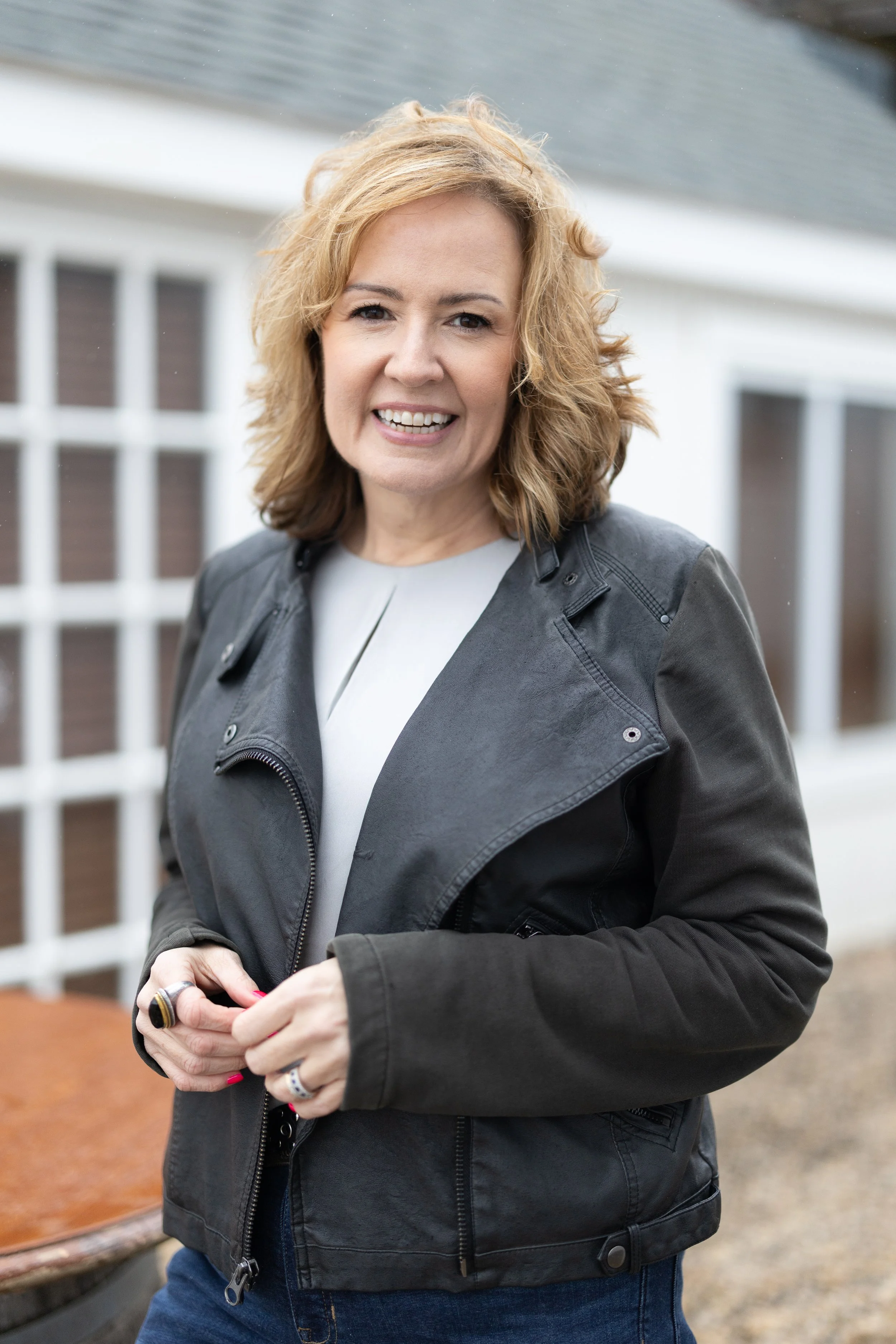A woman with blonde, curly hair smiling at the camera outdoors, wearing a black leather jacket and jeans.