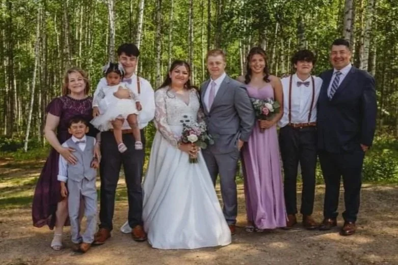 Group of people in formal attire standing outdoors in a wooded area, including a bride in a white wedding dress and a groom in a gray suit, with family members around them.