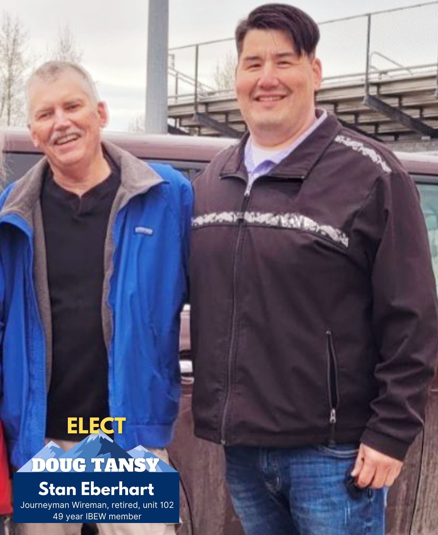 Two men standing outside in front of a vehicle, smiling at the camera. One is wearing a blue jacket, and the other a black jacket. A graphic overlay at the bottom left corner includes the names 'Doug Tansy' and 'Stan Eberhart' along with additional text, indicating a political campaign.