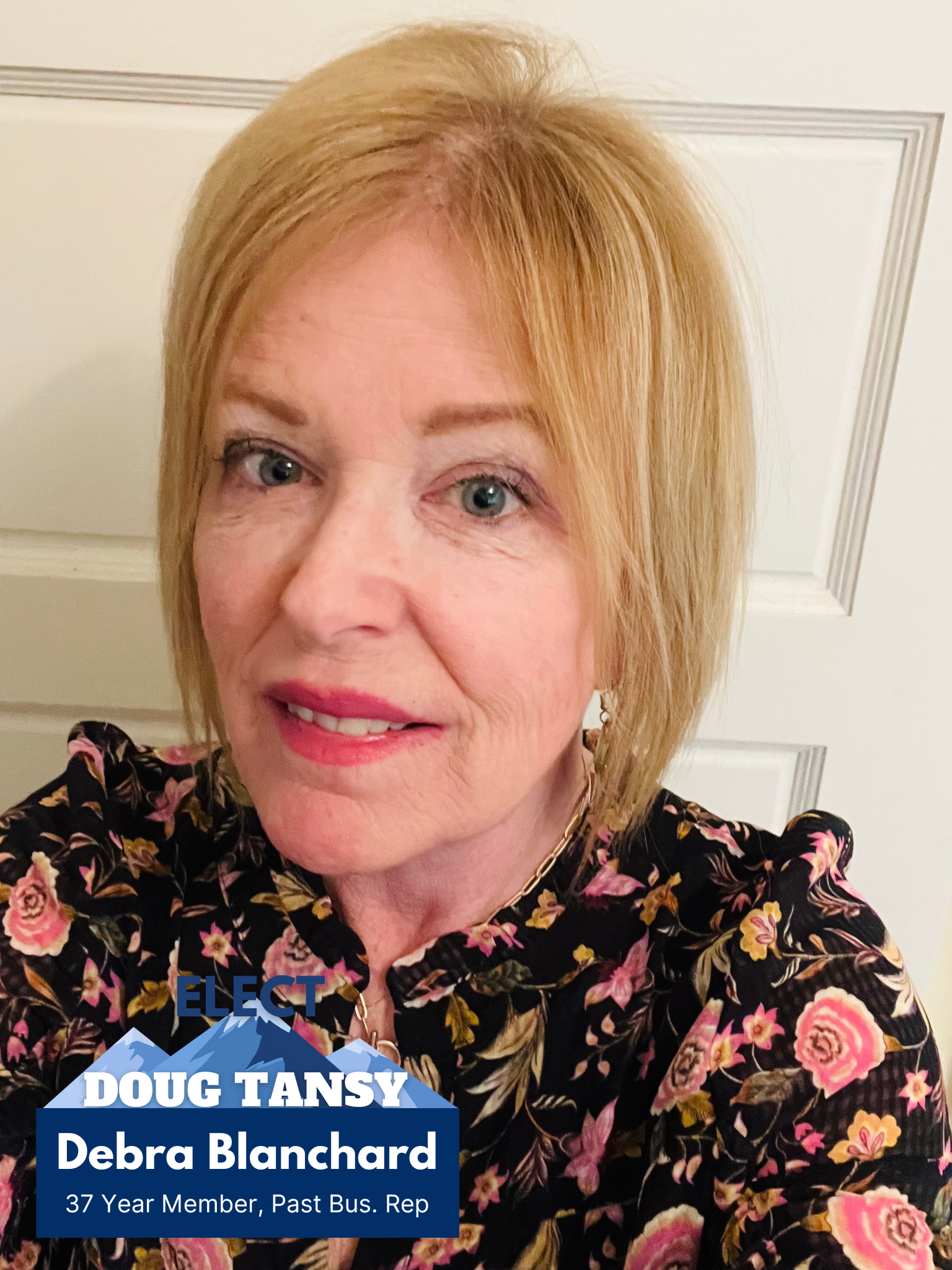 A woman with shoulder-length reddish-blonde hair, blue eyes, and light skin, wearing a black floral blouse, smiling in front of a white door. There is an election campaign graphic with blue mountains and text on the lower left corner that says: 'ELECT DOUG TANSY Debra Blanchard 37 Year Member, Past Bus. Rep.'
