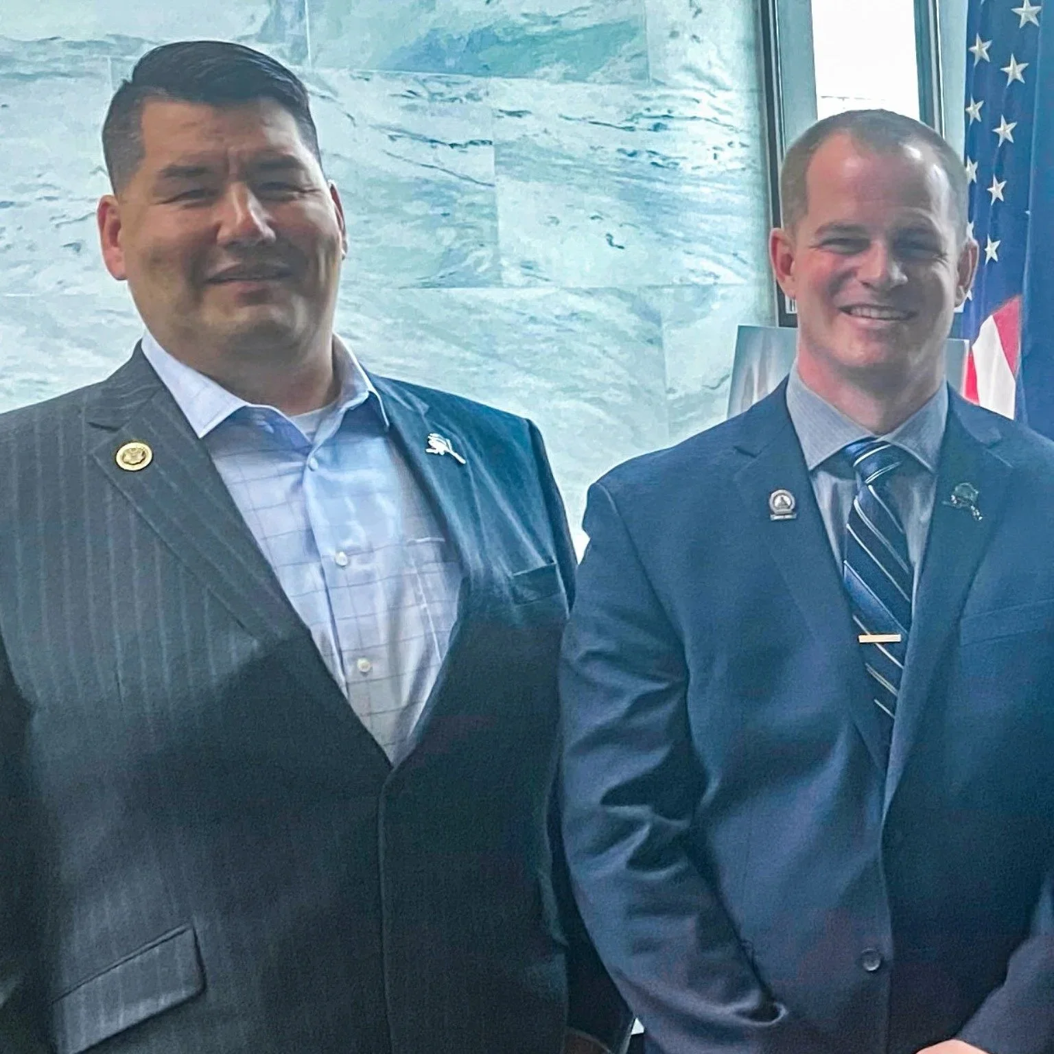 Two men in business suits standing together indoors with an American flag in the background. Both are smiling and wearing lapel pins.