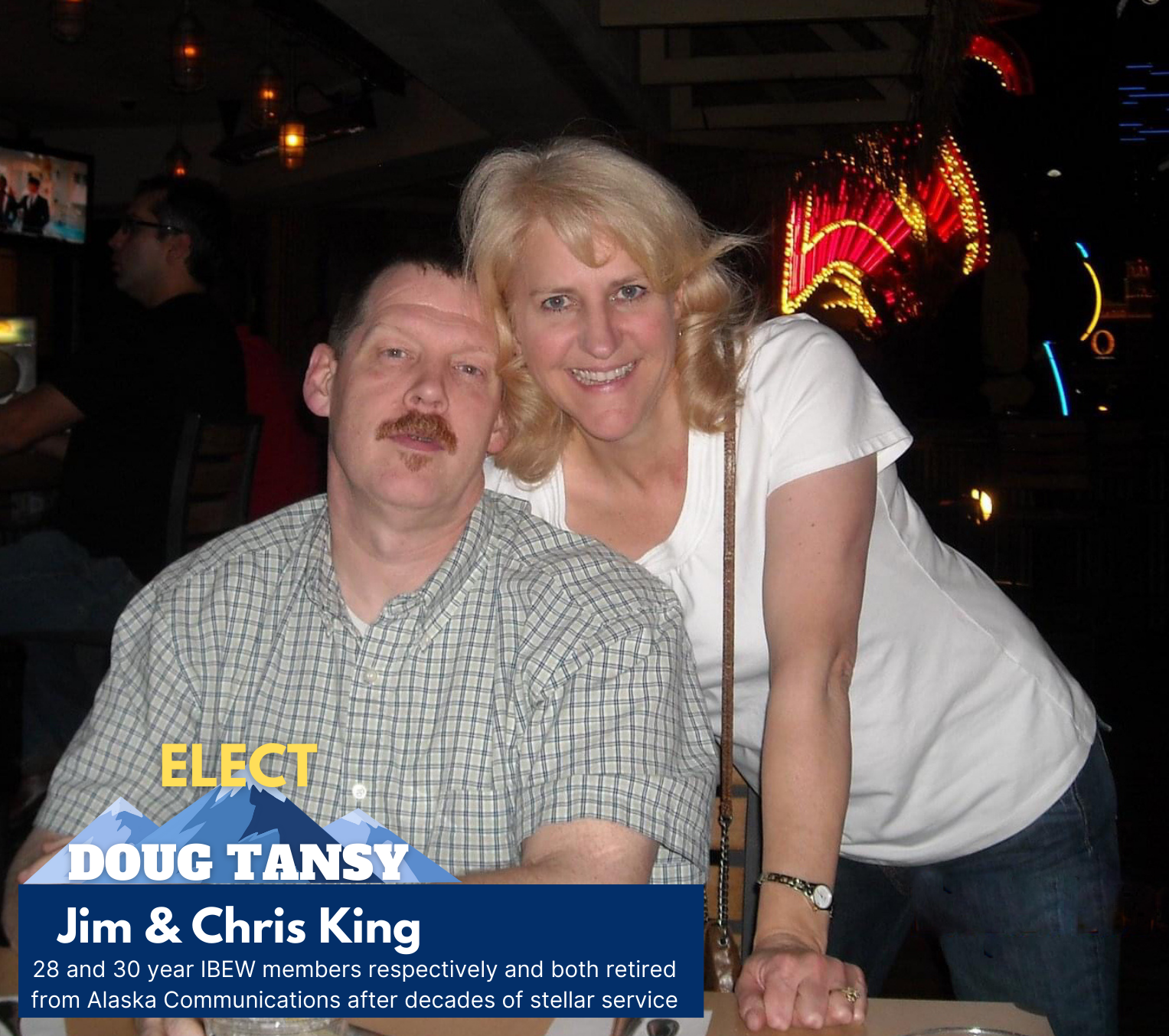 A man and a woman smiling at a social event in a dimly lit venue with colorful neon lights. The man has a mustache and beard, wearing a checkered shirt. The woman has blonde hair, wearing a white t-shirt, and leaning on a table.