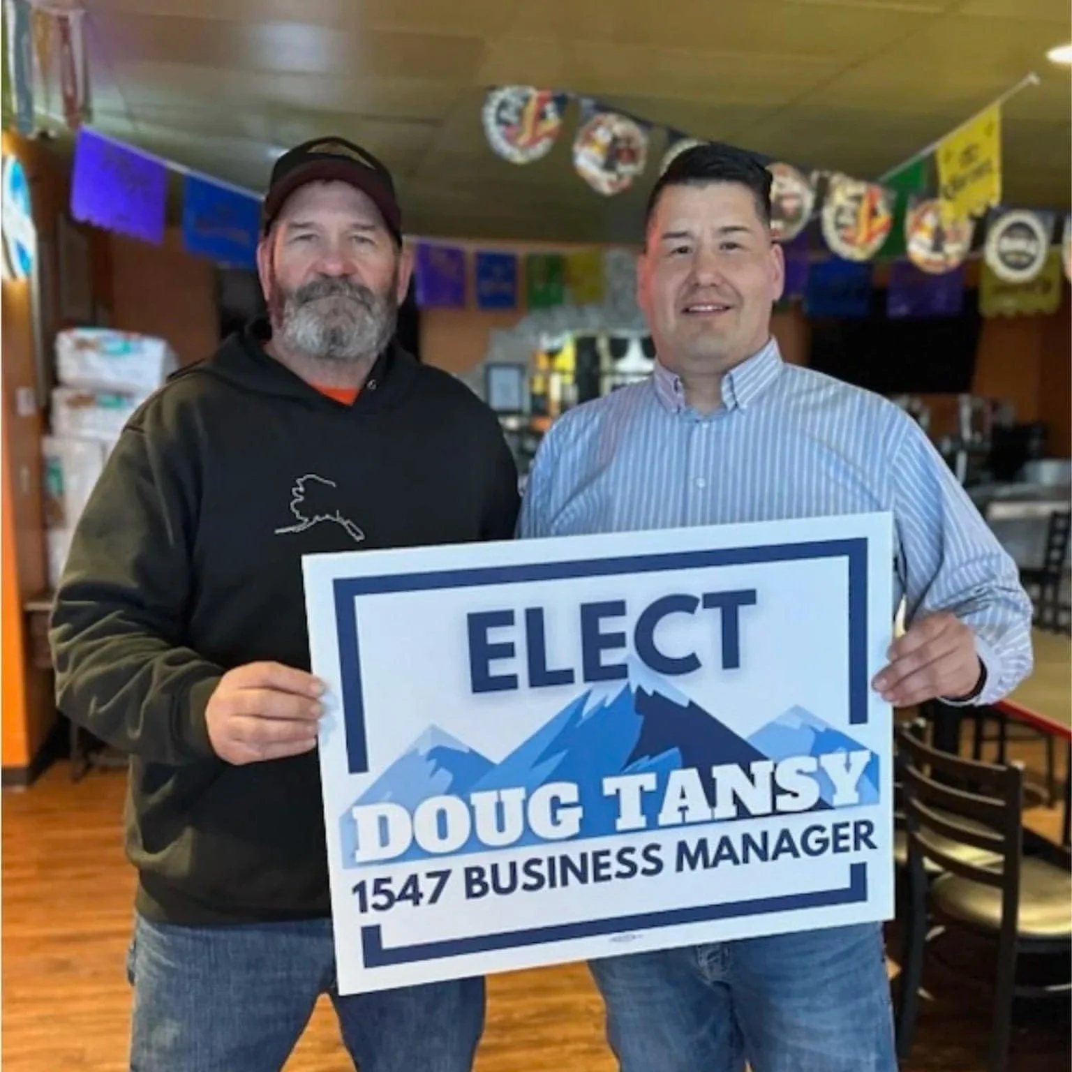 Two men standing indoors, holding a political campaign sign that reads 'Elect Doug Tansy, 1547 Business Manager', with a background of colorful banners and a wooden interior.