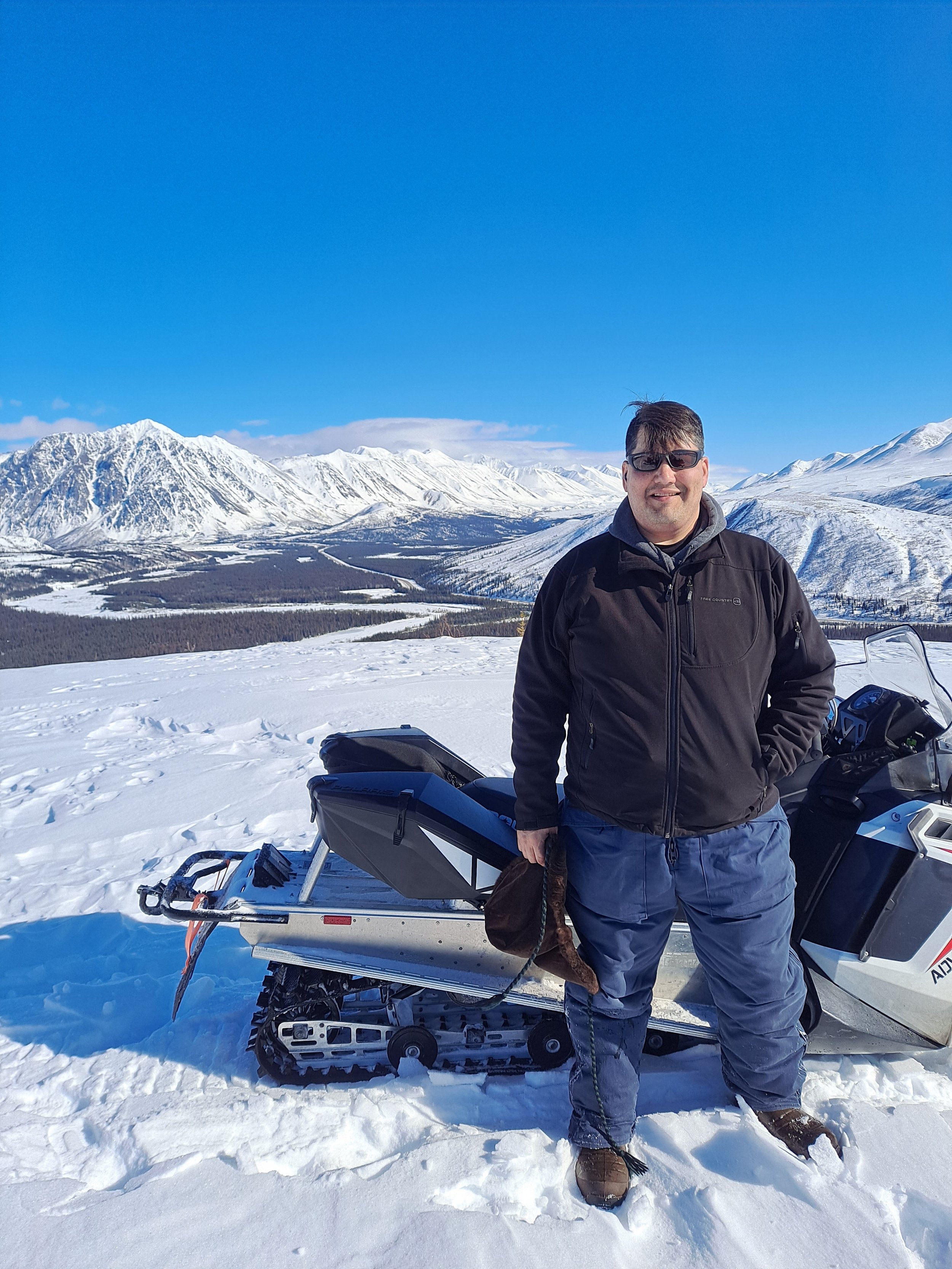 A man in winter clothing standing next to a snowmobile in a snowy landscape with snow-covered mountains in the background under a clear blue sky.