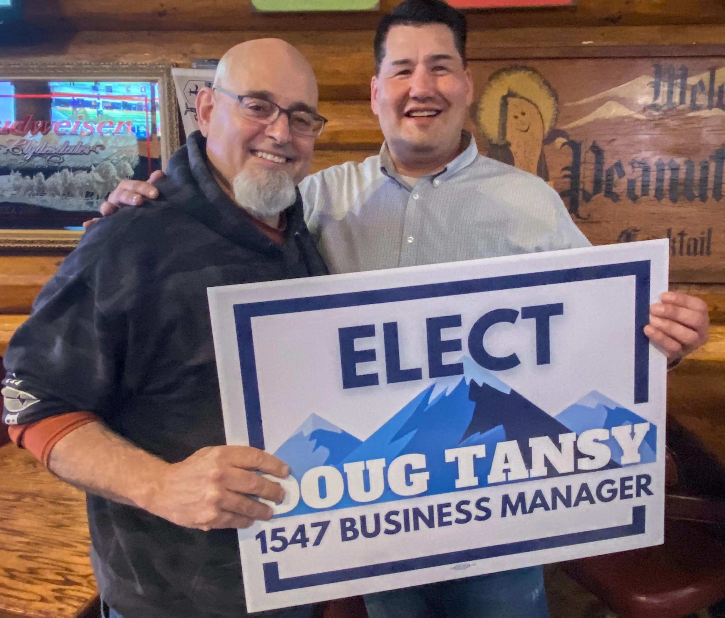 Two men smiling and holding a campaign sign that reads "Elect Doug Tansy, 1547 Business Manager." One man is bald with glasses and a goatee, wearing a black jacket, and the other man has short dark hair, wearing a light-colored shirt. They are standing in a wooden-walled room with signs and a Neon Budweiser sign in the background.