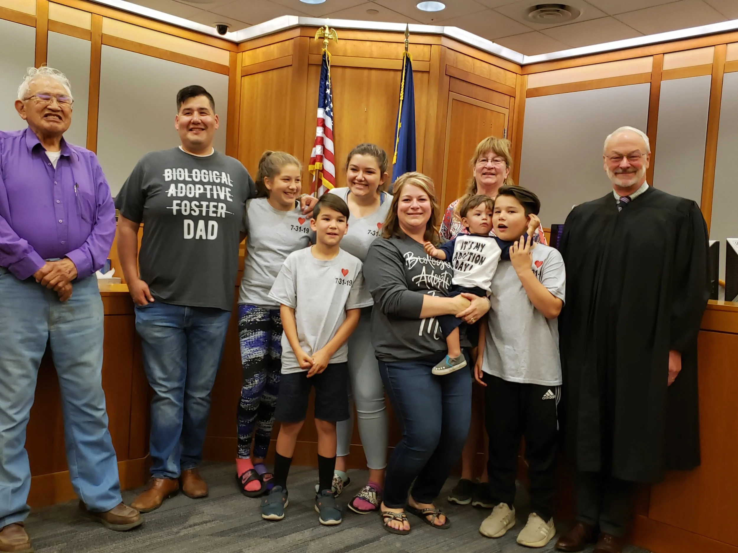 A group of people, including adults and children, standing together in a courtroom or formal setting, celebrating an adoption. Some are wearing shirts with messages about adoption, and there are American and state flags in the background.