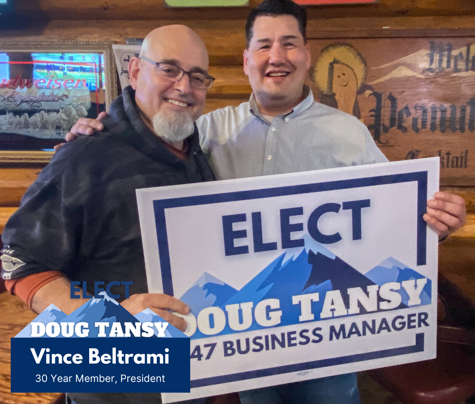 Two men smiling and holding a campaign sign that reads "Elect Doug Tansy for Business Manager, 47". One man is bald with glasses and a beard, wearing a dark jacket, and the other man has dark hair, wearing a light-colored shirt. They are standing in a wooden interior with a neon Budweiser sign in the background.