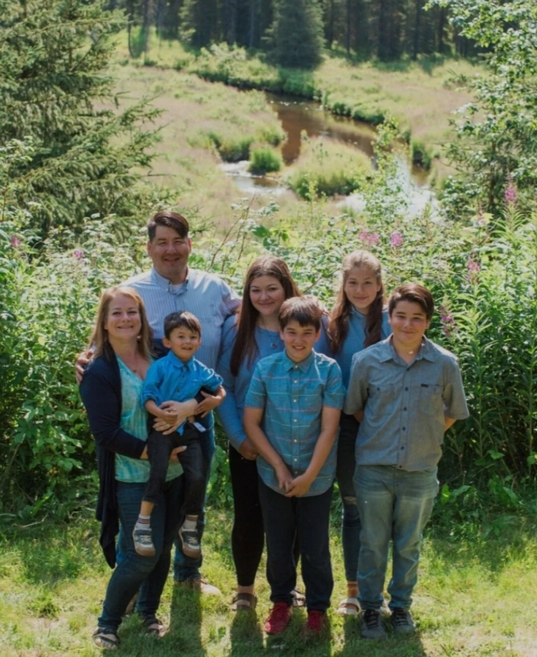 A family of seven outdoors in a lush green area near a small creek, smiling at the camera.