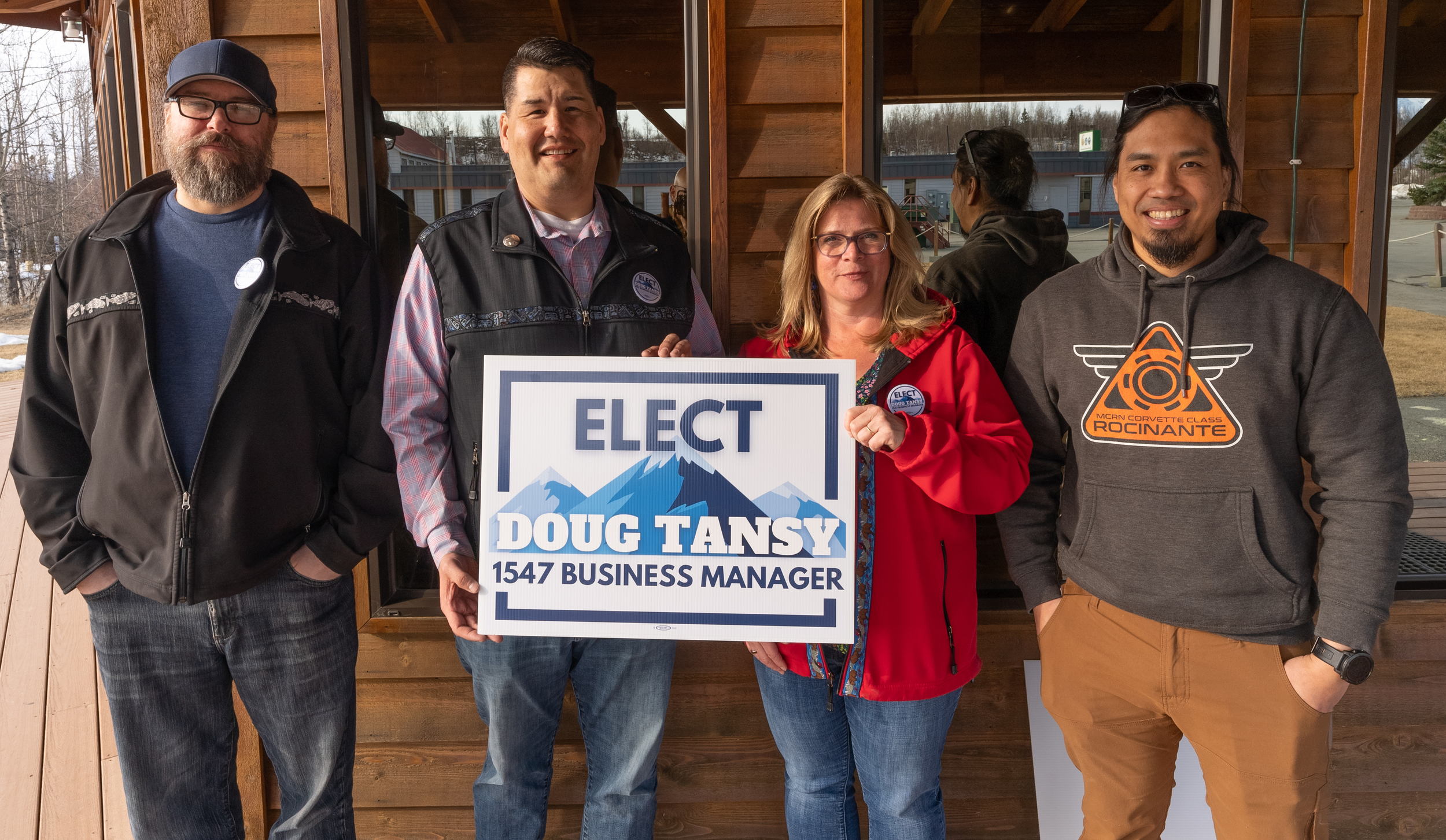 Group of four people standing in front of a wooden building, holding a campaign sign that reads 'ELECT DOUG TANSY 1547 BUSINESS MANAGER.'