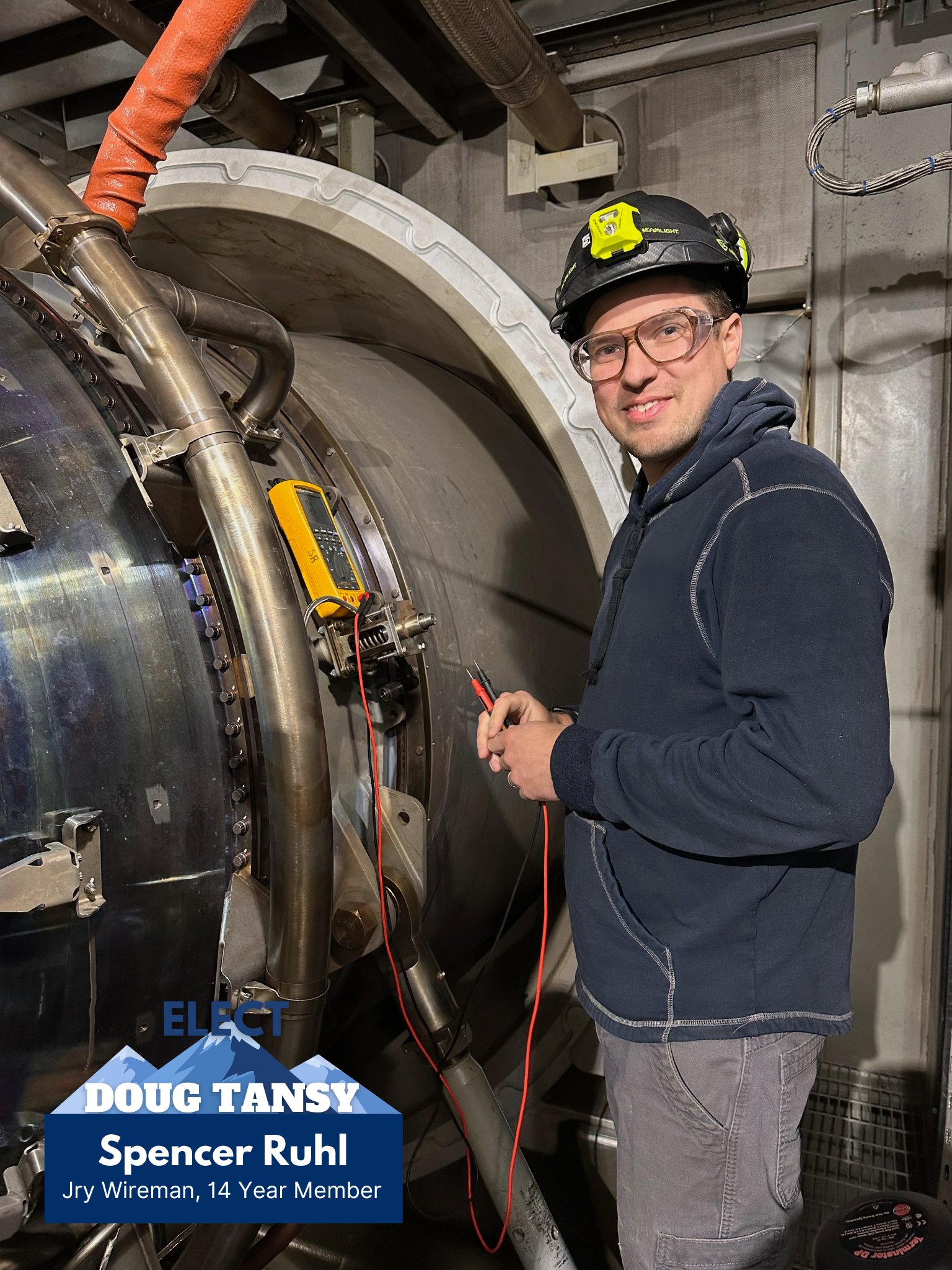 A young man in a black helmet, safety glasses, and dark hoodie standing next to industrial equipment while holding electrical test probes. The equipment appears to be a large machine or reactor within an industrial or laboratory setting.
