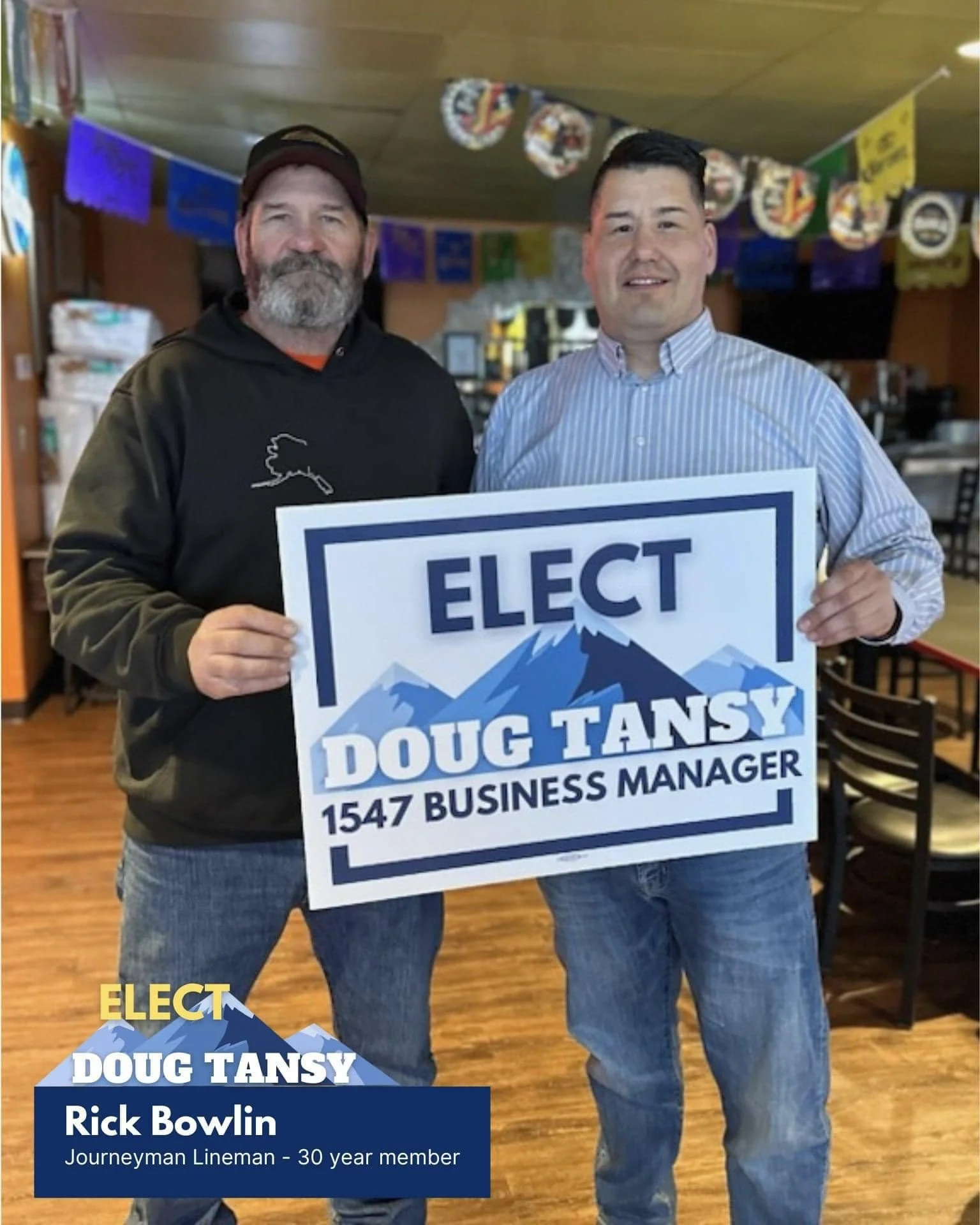 Two men standing indoors, holding a campaign sign for Doug Tansy running for business manager, with colorful decorations hanging in the background.
