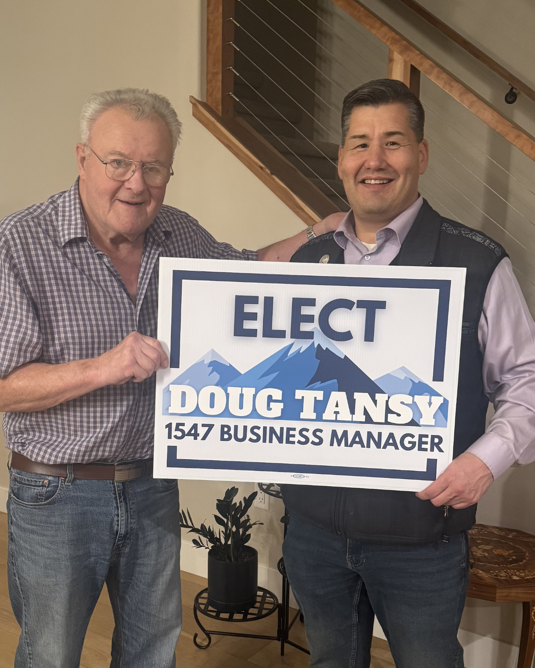 Two men standing indoors, holding a campaign sign that reads 'ELECT Doug Tansy 1547 Business Manager' with mountain graphic, smiling at the camera.