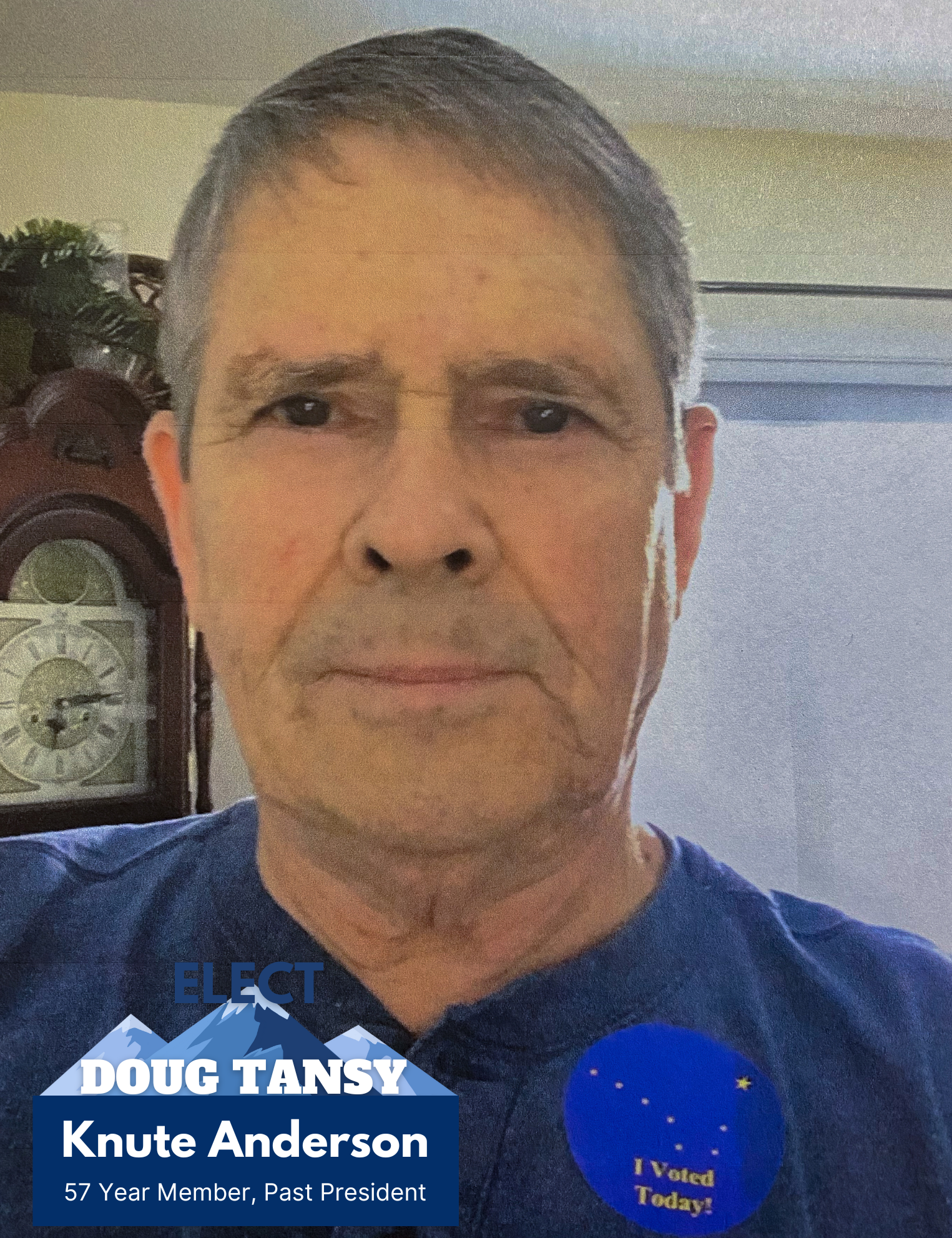 A man with short gray hair wearing a blue shirt and two campaign stickers, one saying 'ELECT Doug TANSY' with mountains and another saying 'I Voted Today!' with stars, standing indoors near a clock.