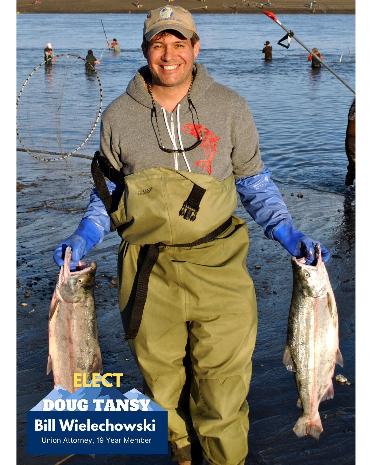 Man smiling at the camera while holding two large fish, one in each hand, near a body of water with several people fishing in the background.