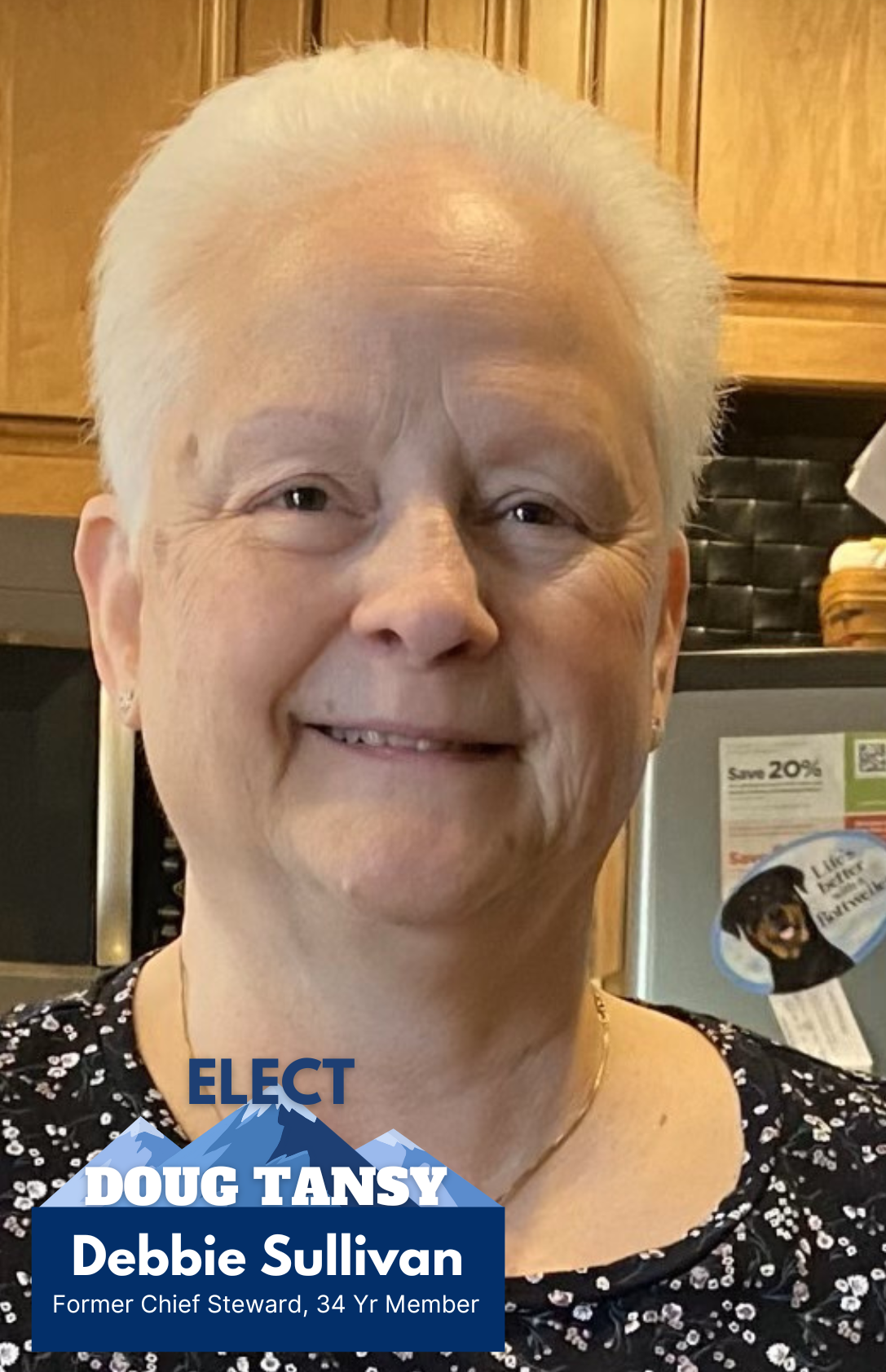 Close-up photo of Debbie Sullivan, an elderly woman with short white hair, smiling, wearing a black floral top, posed indoors in front of wooden cabinets and a refrigerator with notices and a magnet.